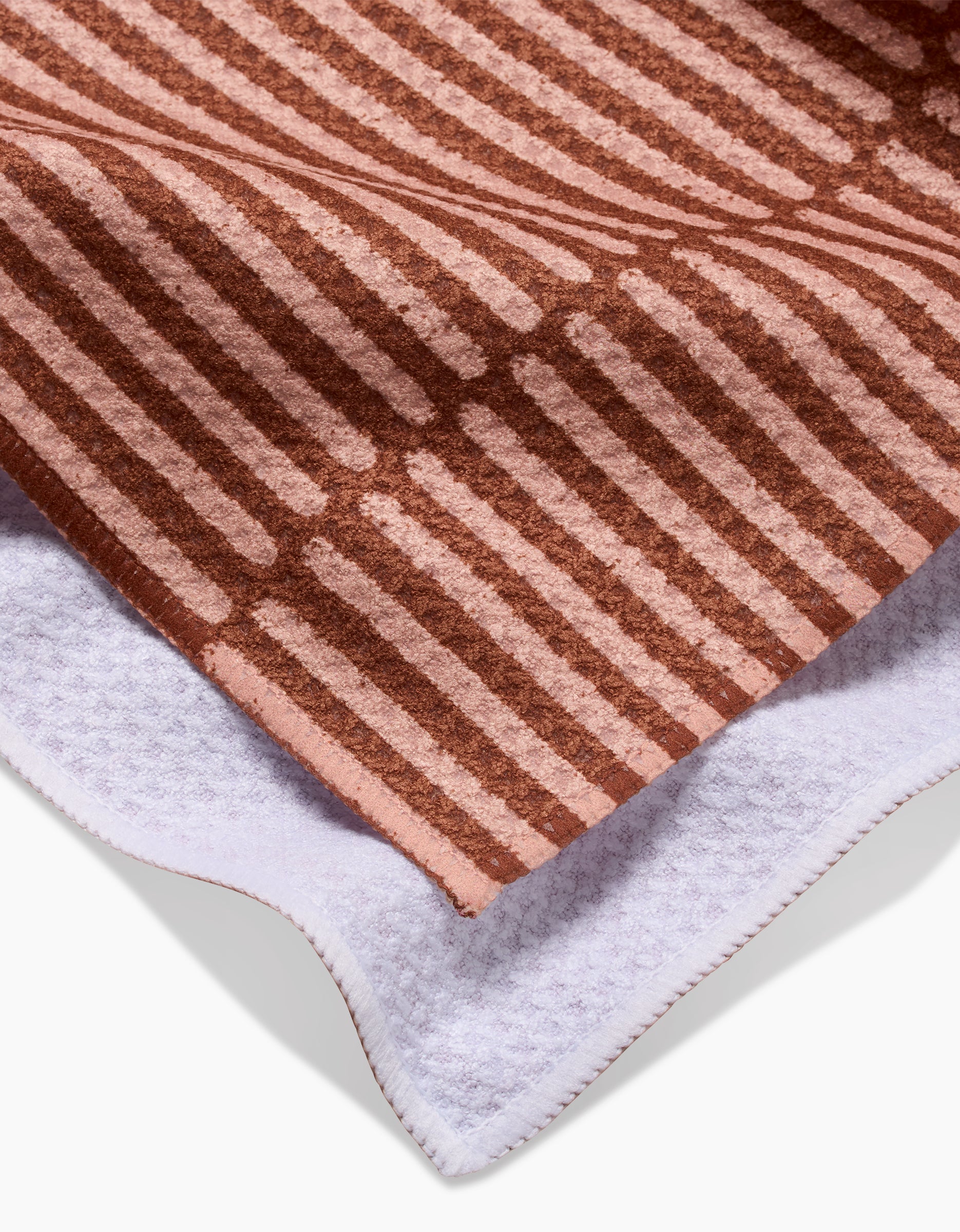 A close-up of a folded Rosa Kichline Textured Stripe Tea Towel shows a brown and pink striped pattern on one side and a plain white ultra-absorbent Geoweave fabric surface on the other.