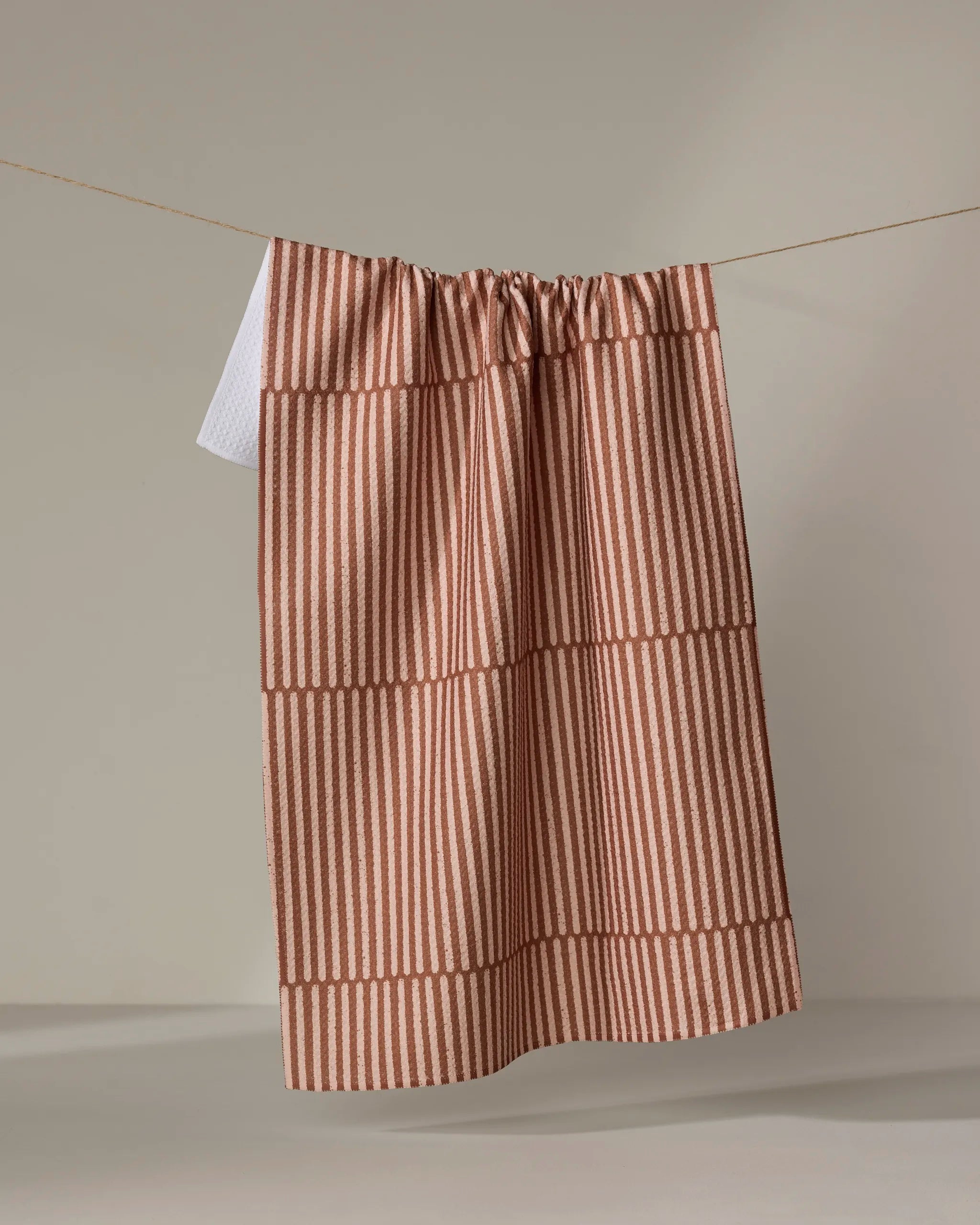 The Rosa Kichline Textured Stripe recycled kitchen towel in rust, featuring vertical and horizontal stripes, hangs on a clothesline against a plain beige backdrop.