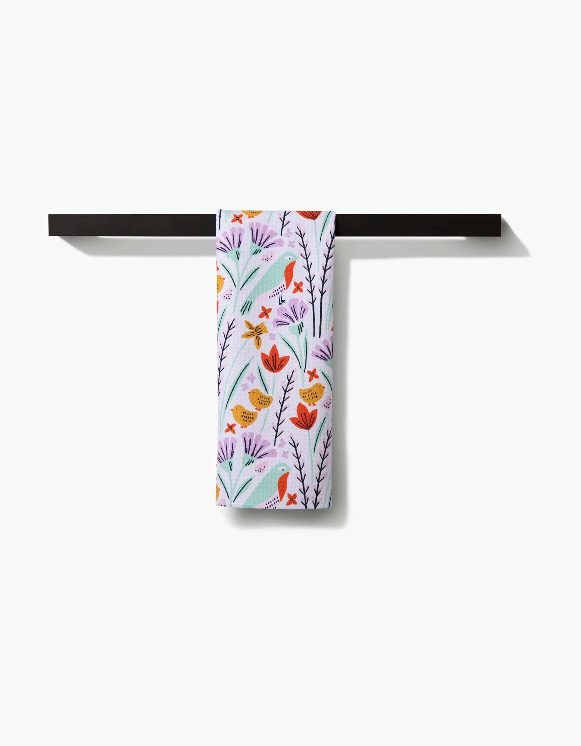 The Amy MacCready “Thaddeus” tea towel, featuring a vibrant floral and bird design, hangs neatly on a black horizontal towel bar against a white background.
