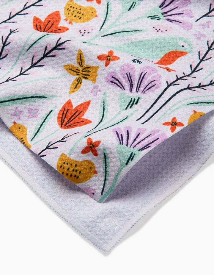 A close-up of the Thaddeus tea towel by Amy MacCready, featuring a colorful botanical print with flowers, leaves, and small birds. A folded corner shows the ultra-absorbent textured underside.