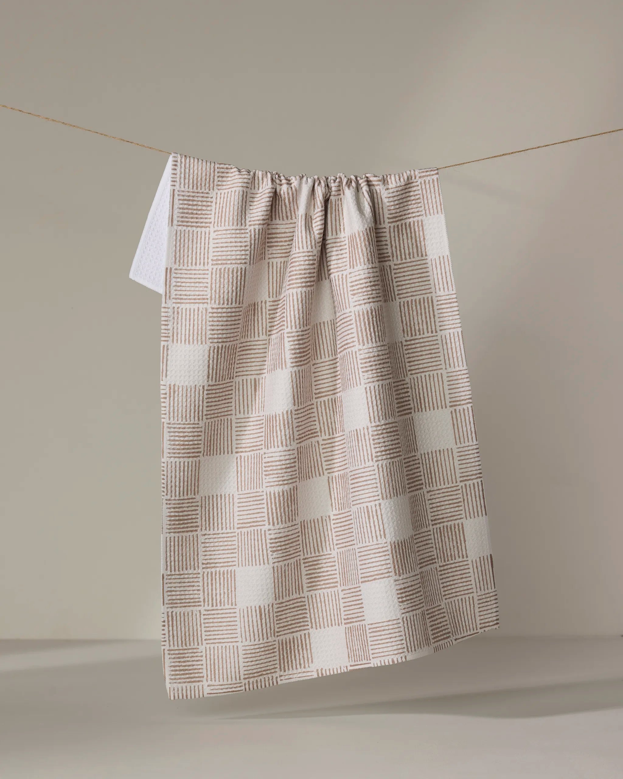 The Thatcher tea towel by Studio Jeannot, featuring a beige and white checkered pattern and crafted from recycled materials, hangs on a string against a neutral backdrop, displaying soft folds and gentle shadows.