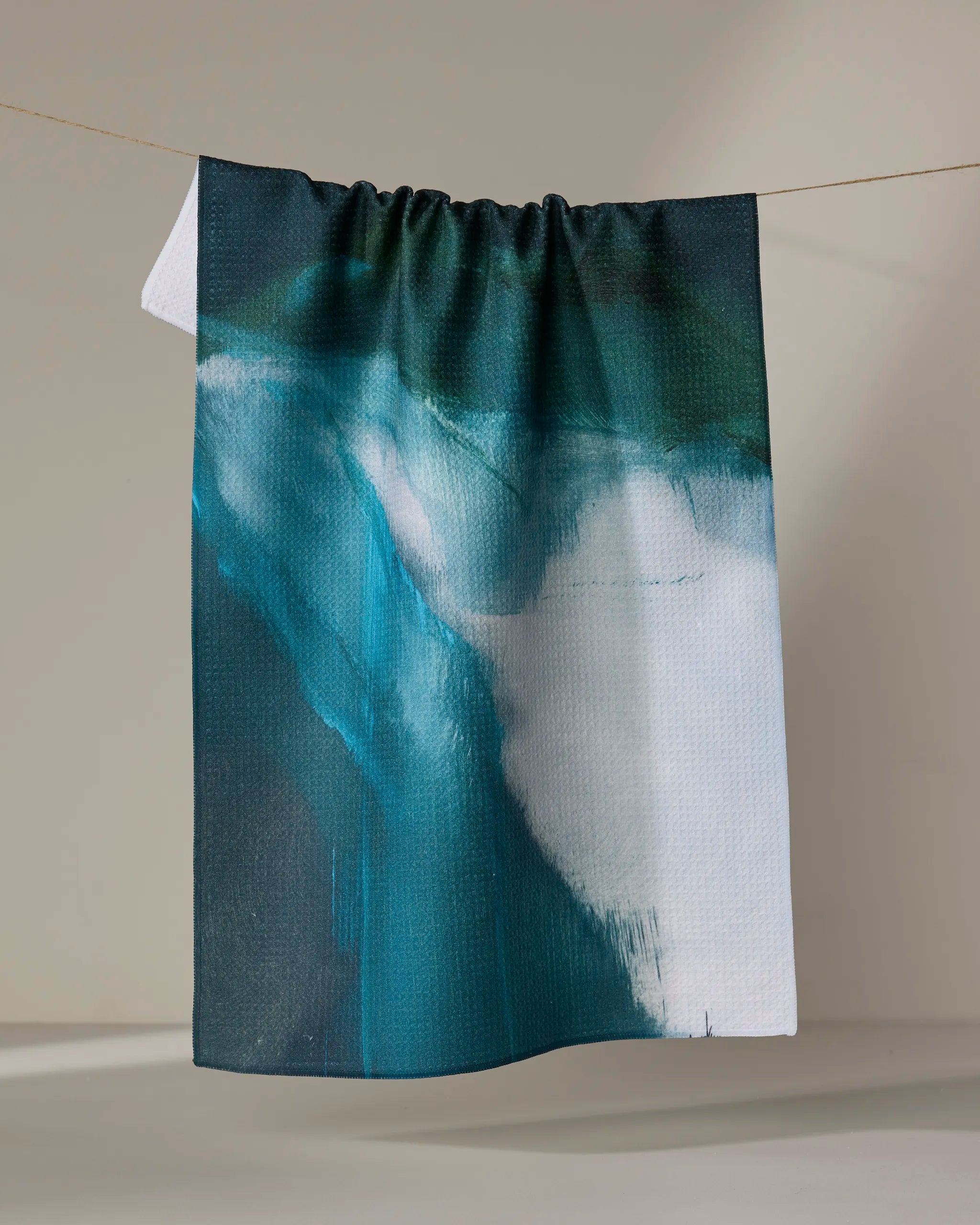 The Air We Breathe" tea towel by Jennifer Janesko, featuring an abstract design in dark green, teal, and white, hangs on a thin line against a plain beige background.