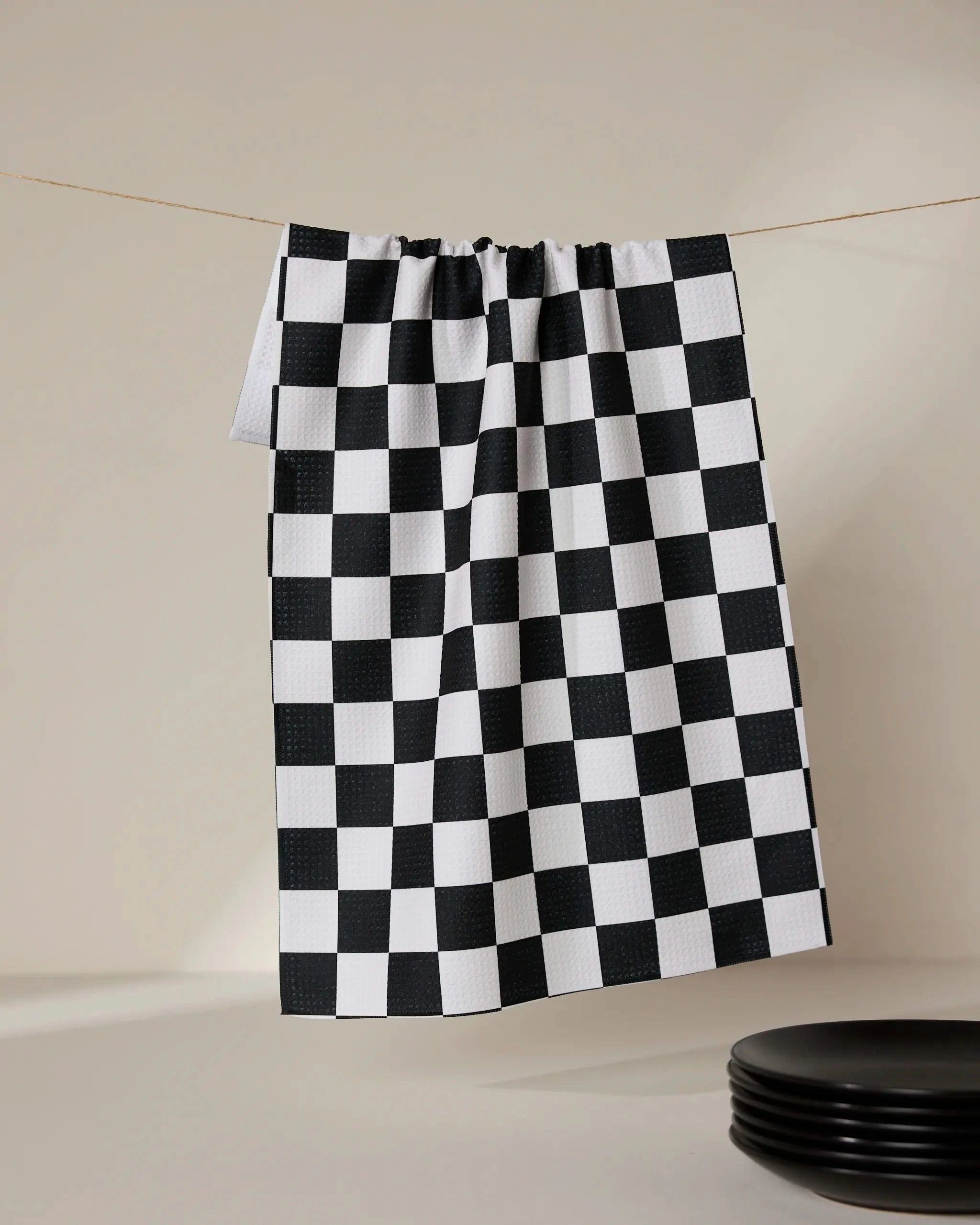 The Classic tea towel by GEOMETRY, made from recycled materials in a black and white checkered pattern, hangs from a clothesline against a neutral background with black plates stacked in the lower right corner.