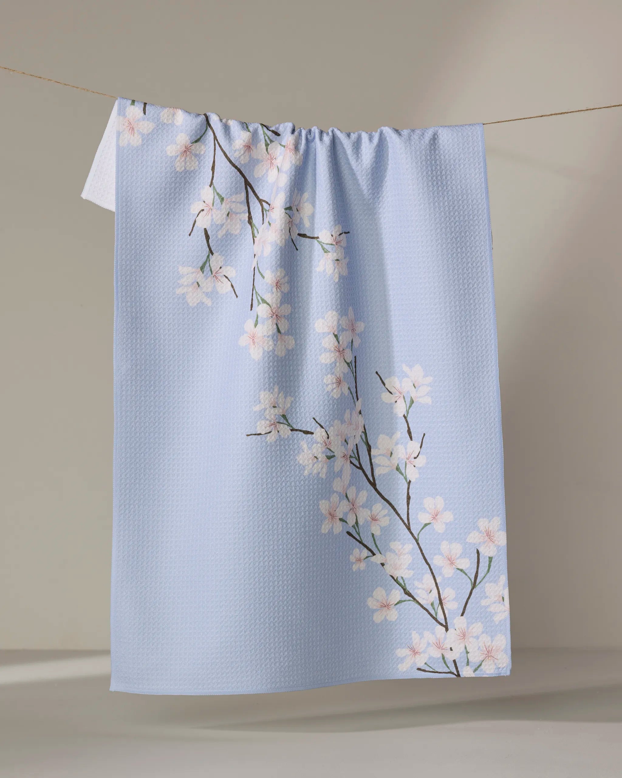 The Eve by Becki Owens is a light blue Geoweave tea towel with white and pink cherry blossom branches in a textured pattern, displayed hanging on a clothesline against a neutral backdrop.