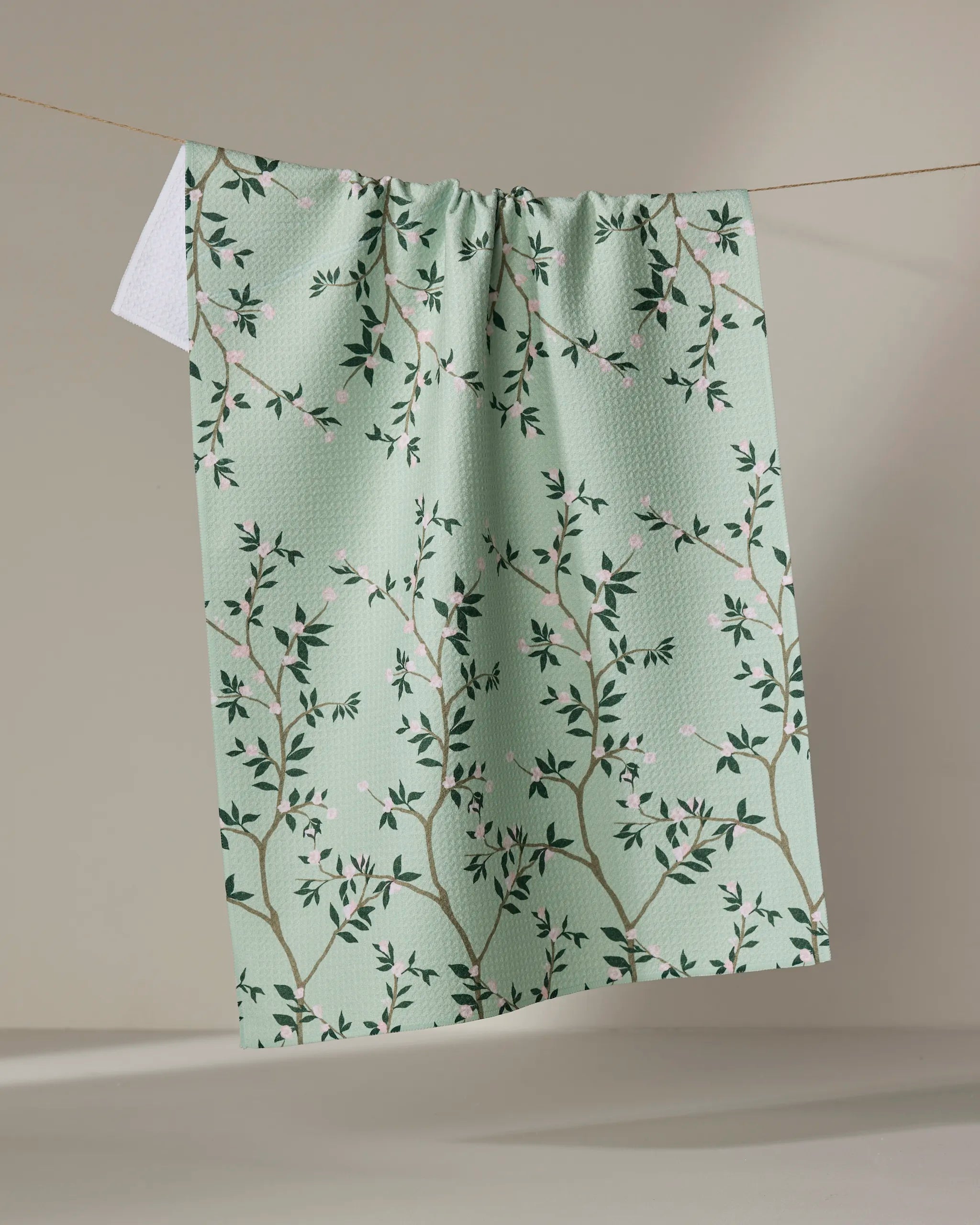 The Lily by Becki Owens is a light green kitchen tea towel featuring a branch and leaf pattern with pink flowers. Made from recycled materials, it’s an eco-friendly and charming accent for your home.