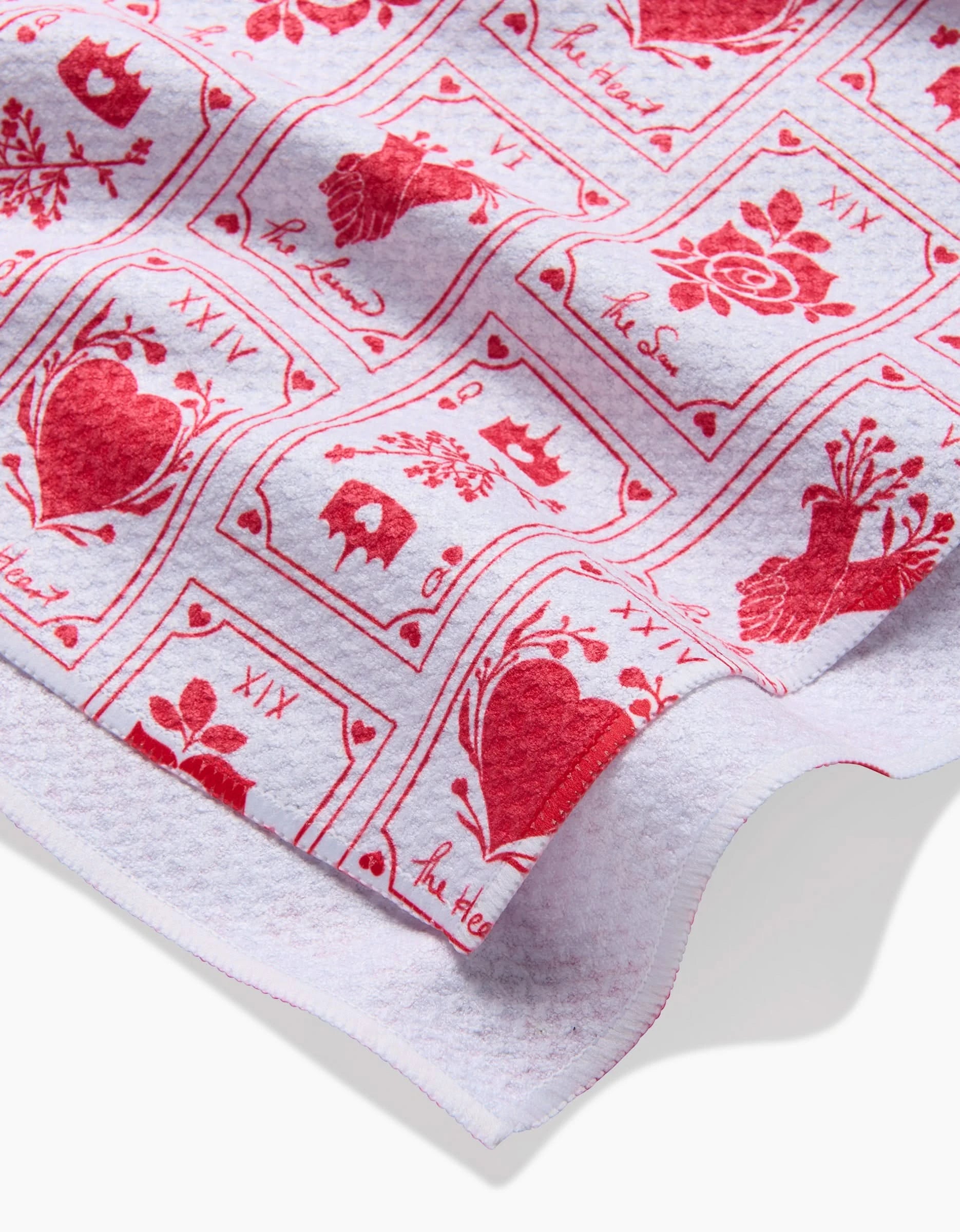 A close-up of The Lovers tea towel by Danelys Sidron, made from recycled materials. It features a red heart, flower, and bird print in squares with Ah, Heart and XIX text on white fabric; edge detail shown.