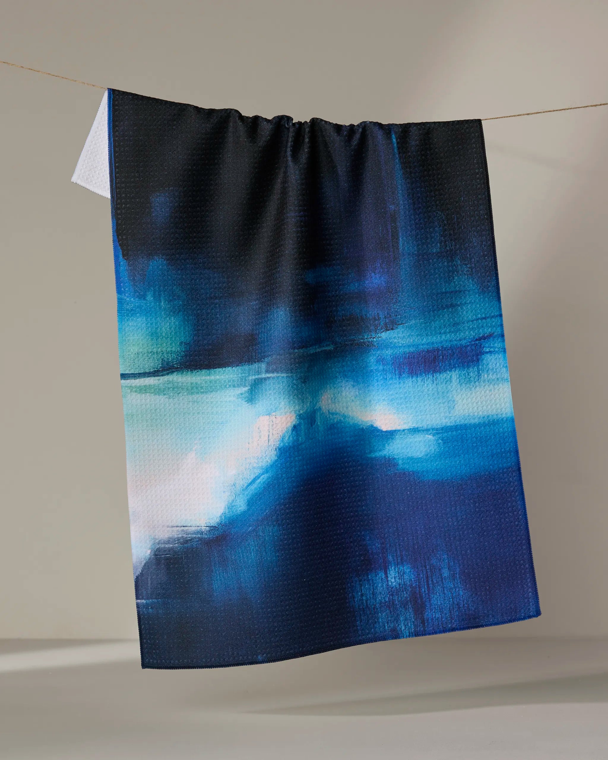 The Ocean Between Us" tea towel by Jennifer Janesko, featuring an abstract blue, black, and white pattern, hangs on a string against a plain beige background.