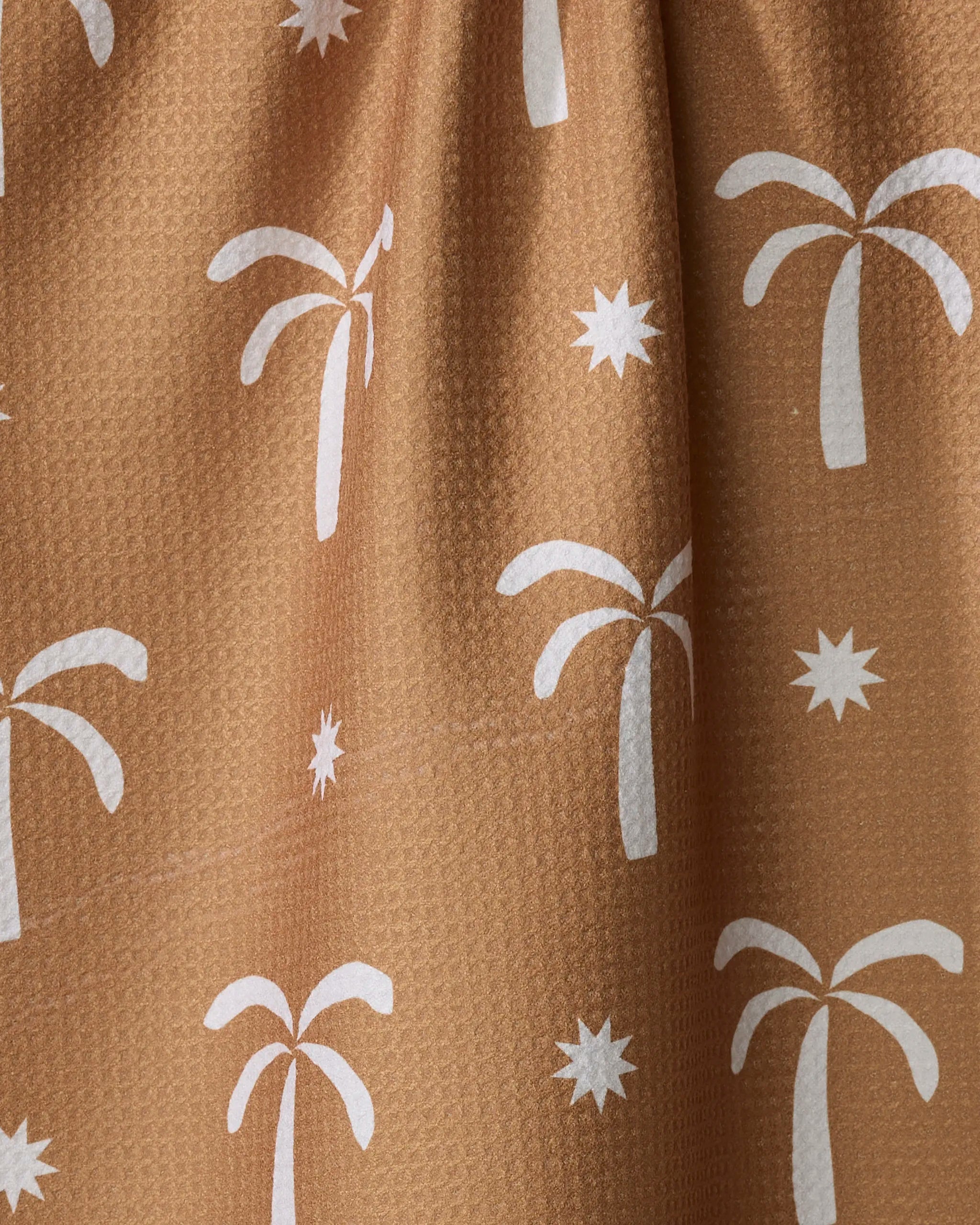 A close-up of The Perfect Palm tea towel by Elizabeth Edwards, made from ultra-absorbent brown textured fabric and adorned with a white palm tree and starburst pattern.