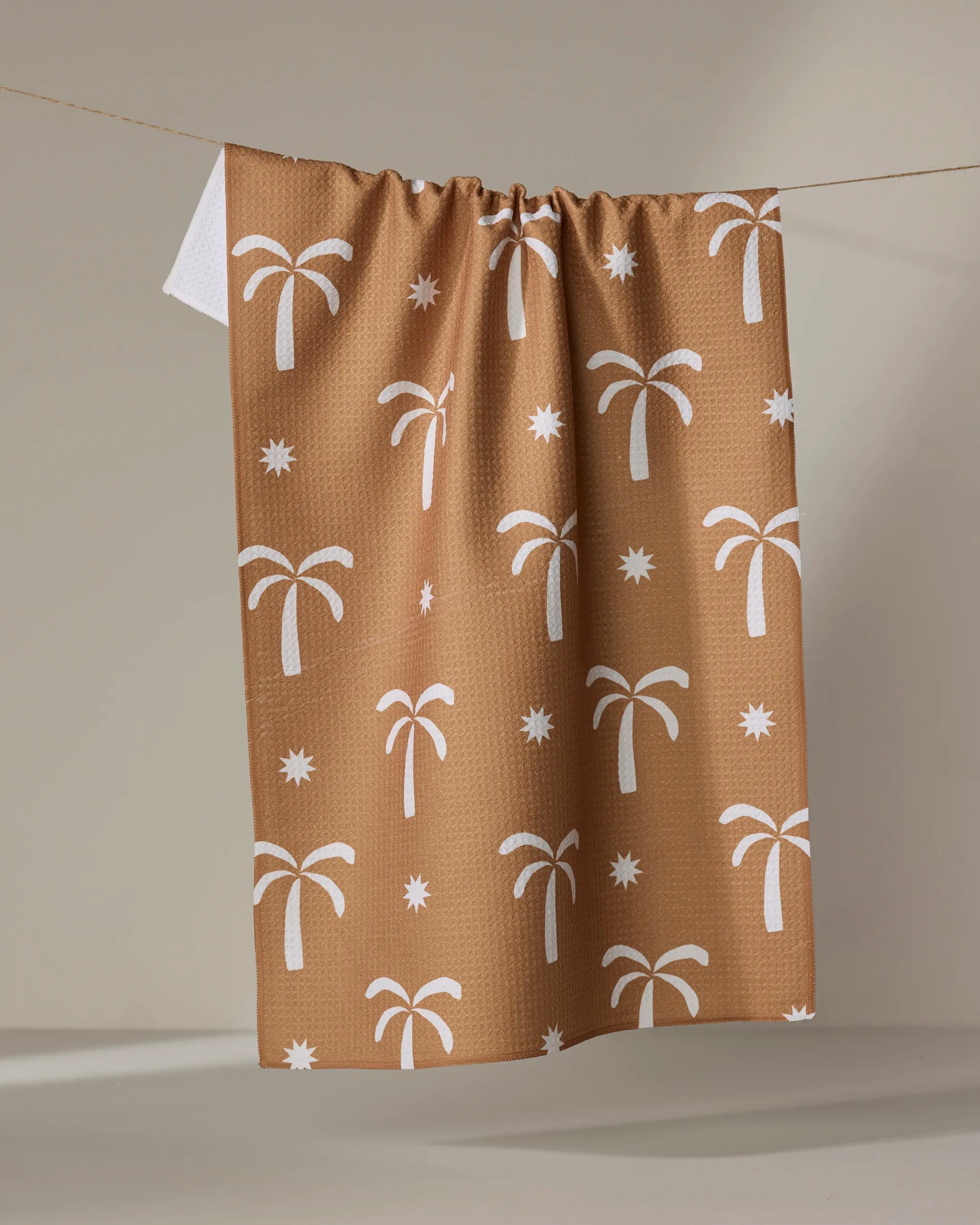 The Perfect Palm tea towel by Elizabeth Edwards features a white palm tree and star pattern on brown. Made from ultra-absorbent material, it’s both stylish and practical—a charming addition to any home.