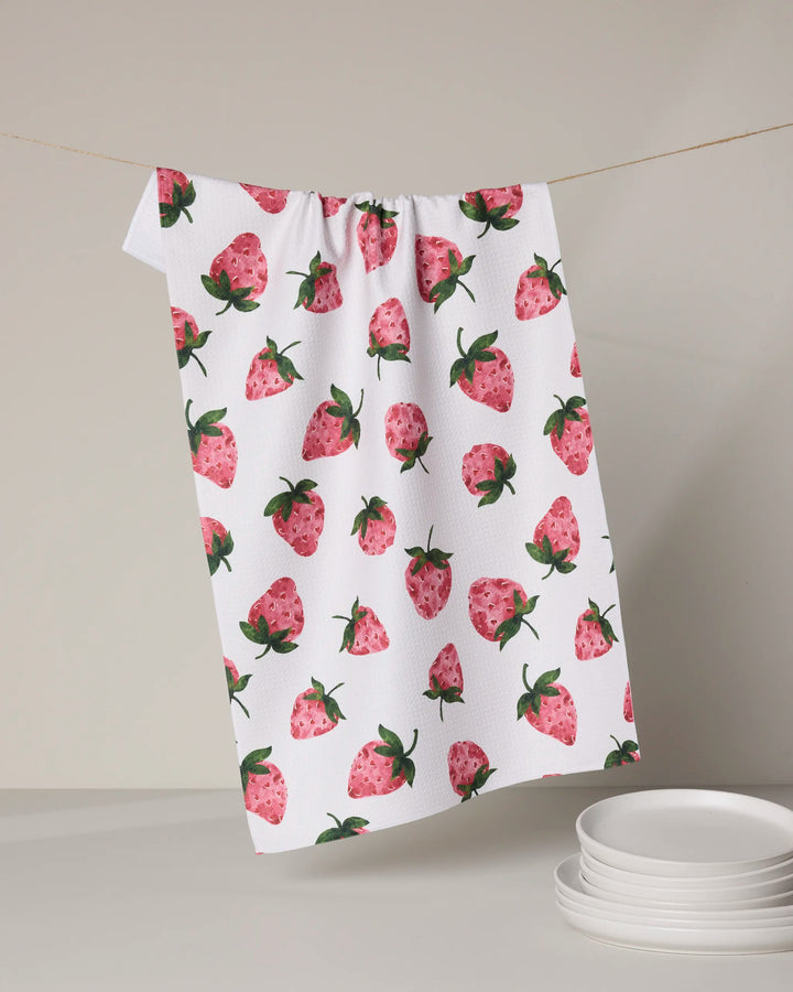 The Sweetest tea towel by Geometry, featuring an ultra-absorbent white fabric with pink strawberry patterns, hangs above a stack of white plates against a light backdrop.