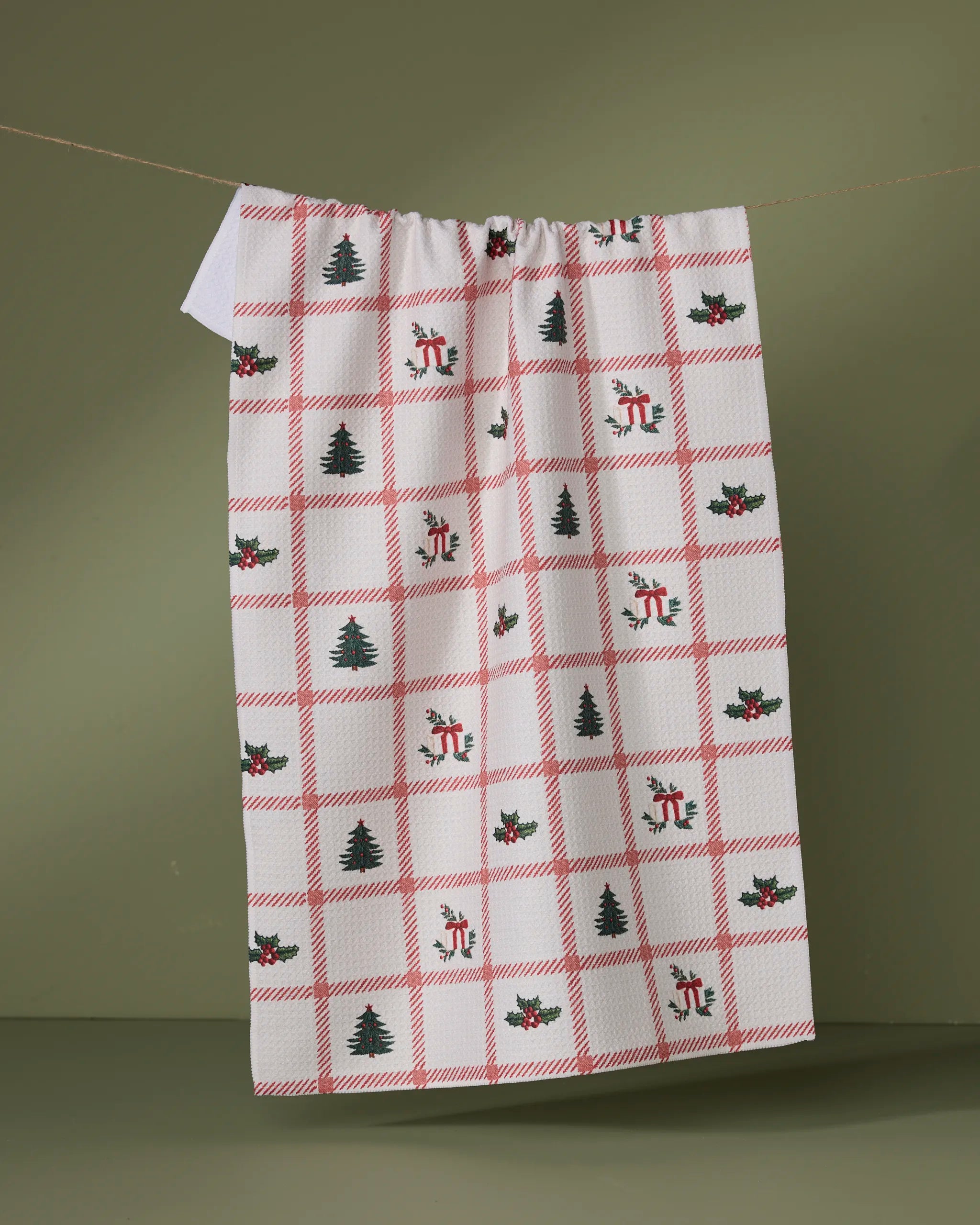 The Geometry Tidings tea towel, featuring quick-dry fabric, red plaid lines, and festive Christmas designs—trees, holly, and bells—hangs against a plain olive green background.