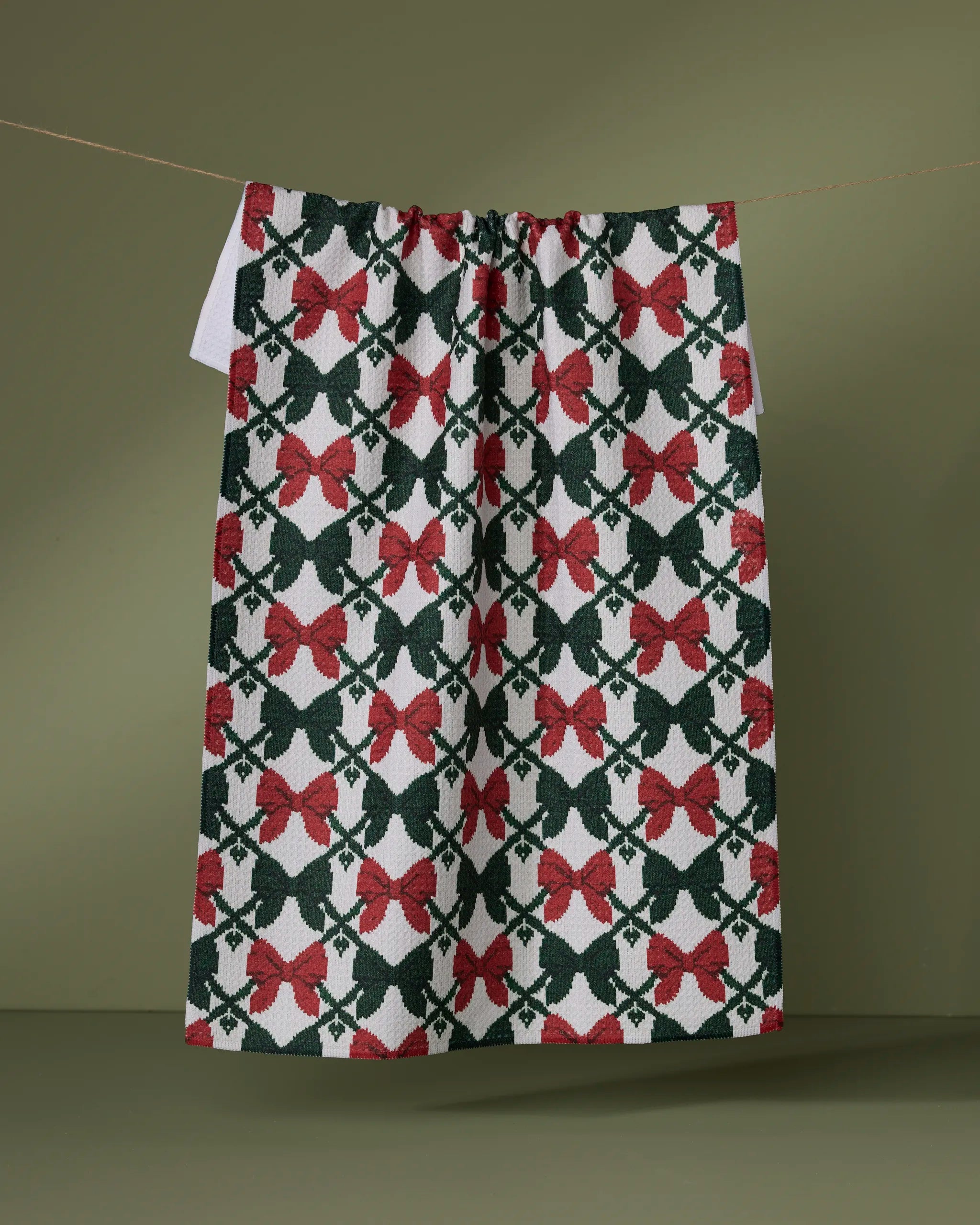 The "Tied" tea towel by Geometry, featuring red bows and green holly leaves, hangs on a string against an olive green background, bringing festive holiday charm to your kitchen.