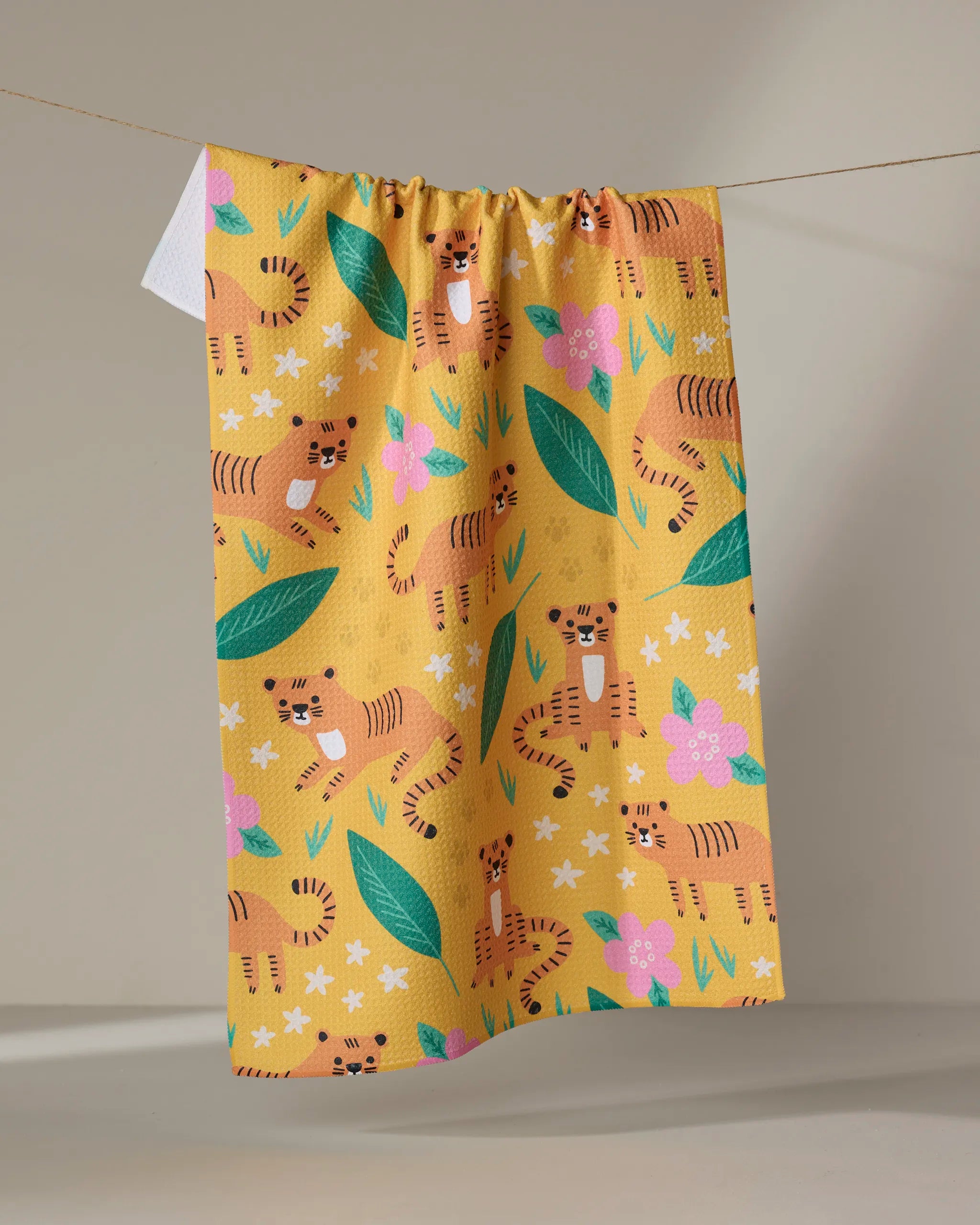 The "Tigers" kitchen towel by Genna Blackburn features cartoon tigers, green leaves, and pink and white flowers in a playful, colorful yellow design.