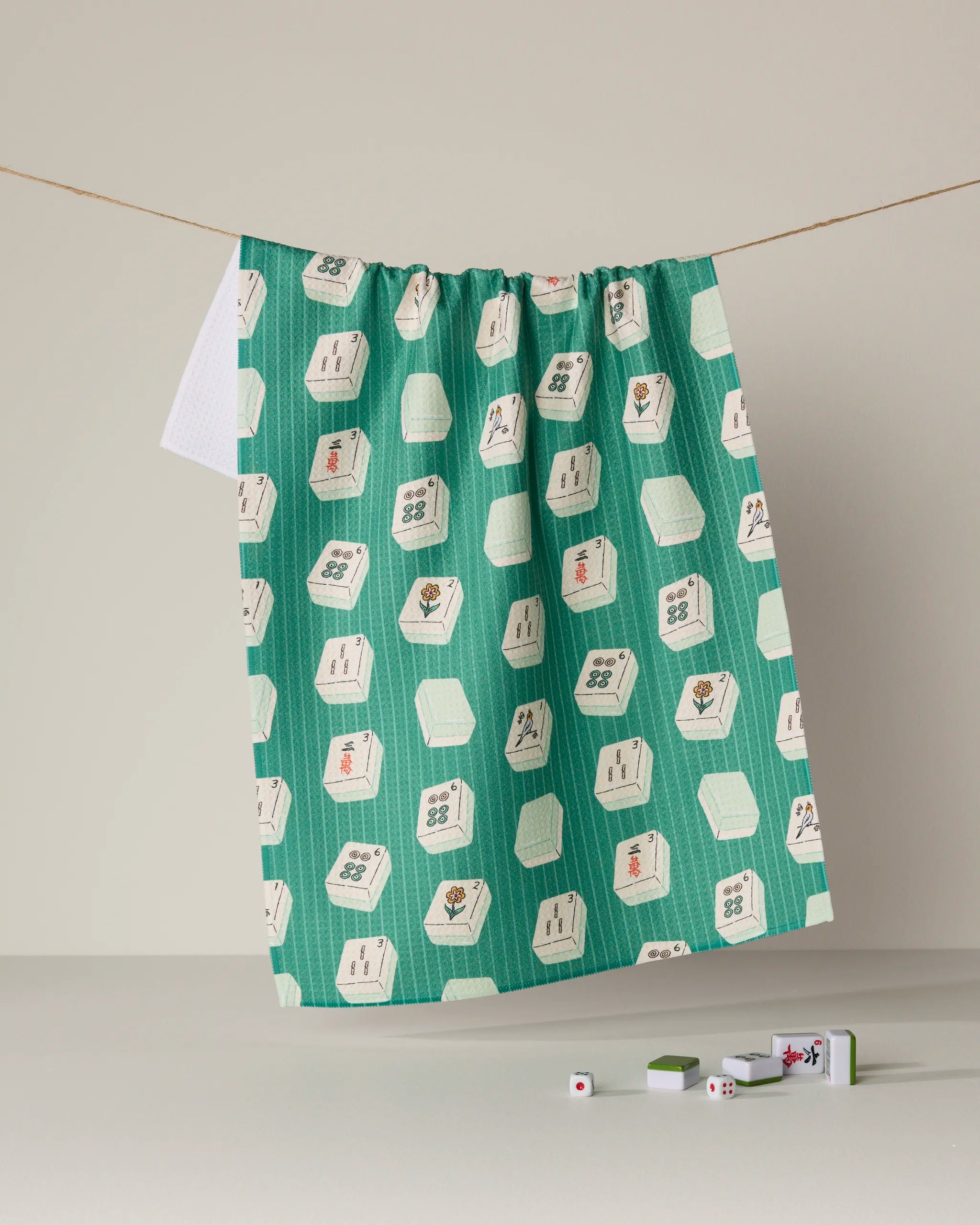 A Geometry "Tile Flow" ultra-absorbent towel—featuring green mahjong tile patterns—hangs on a line, while several mahjong tiles rest below on a light surface against a neutral beige background.