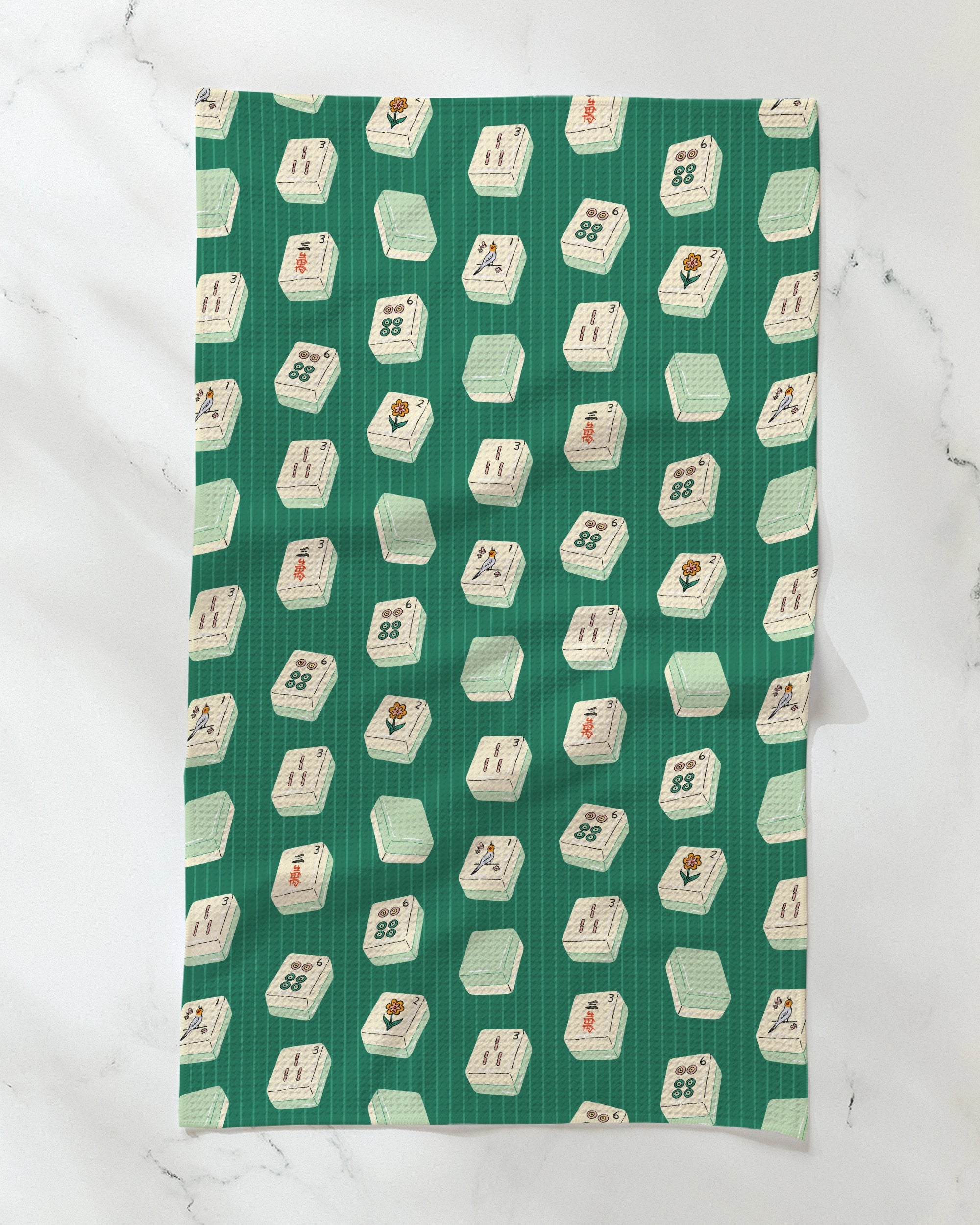 The Geometry Tile Flow tea towel, crafted from recycled materials and featuring a repeating pattern of white mahjong tiles with various symbols and characters on a green cloth, is spread out on a white marble surface. This ultra-absorbent, eco-friendly kitchen accessory combines unique style with practical functionality.