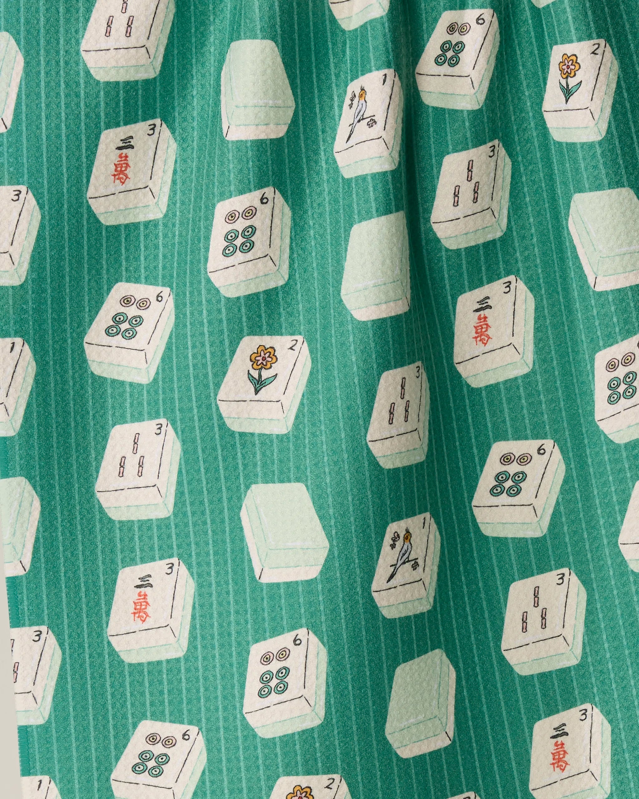 The Geometry Tile Flow tea towel features a green fabric adorned with neat rows of white mahjong tiles, each displaying an array of symbols, dots, and flowers.