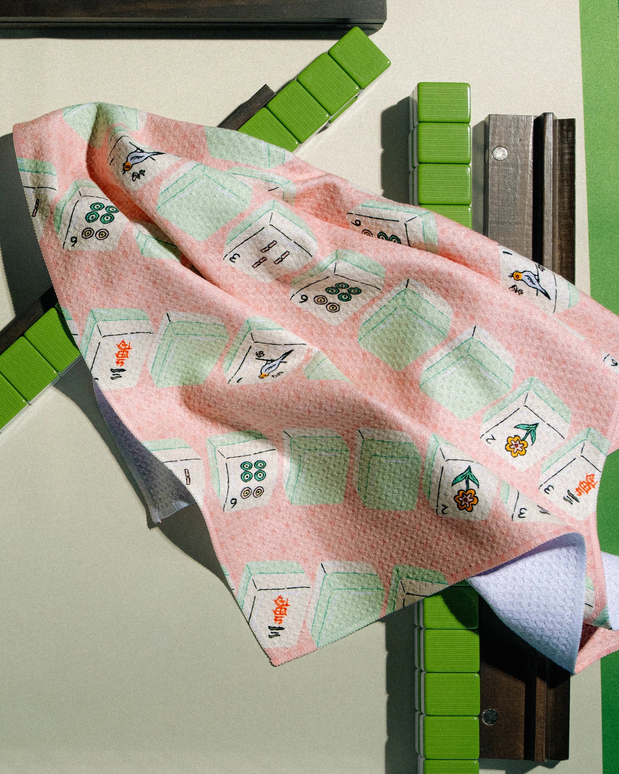 A Geometry "Tile Parade" tea towel—featuring a vibrant pink and white geometric pattern with green and orange accents—is draped over a structure of brown planks and green rectangular blocks, set against a neutral background. Crafted from recycled materials, this ultra-absorbent cloth from Geometry brings both sustainable style and functionality to any space.