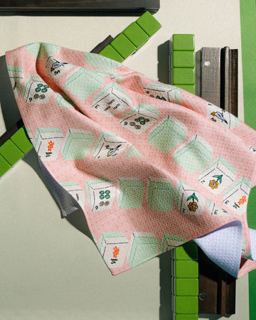 A Geometry "Tile Parade" tea towel—featuring a vibrant pink and white geometric pattern with green and orange accents—is draped over a structure of brown planks and green rectangular blocks, set against a neutral background. Crafted from recycled materials, this ultra-absorbent cloth from Geometry brings both sustainable style and functionality to any space.