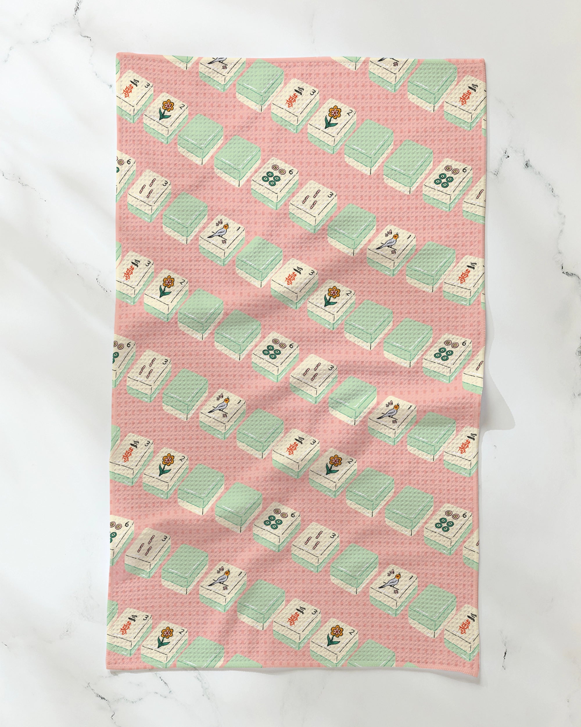 The Geometry Tile Parade tea towel features a pink background decorated with a repeating pattern of light green Mahjong tiles and delicate flowers. Made from ultra-absorbent, recycled materials, it is shown displayed on a white marble surface.