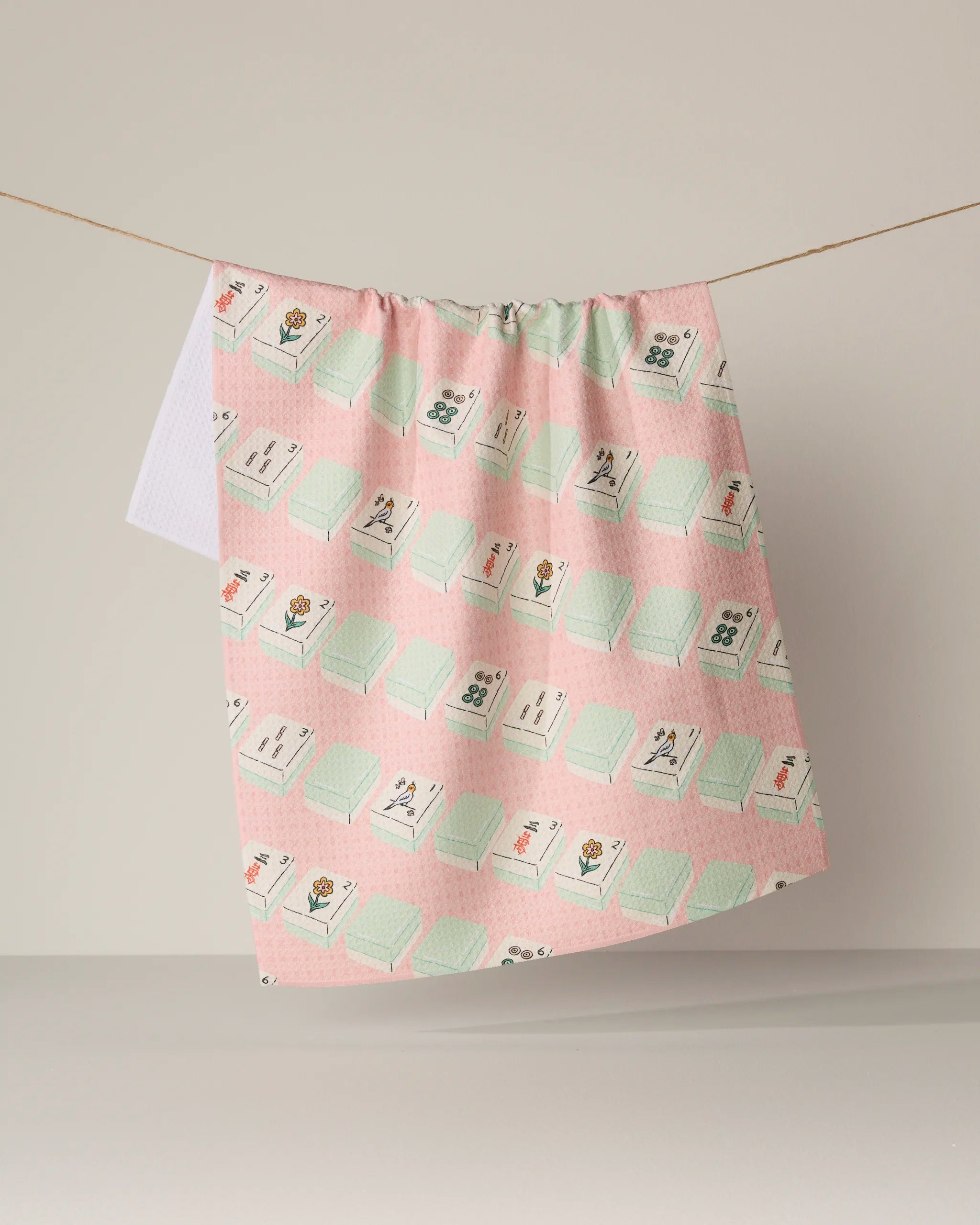 The Geometry "Tile Parade" kitchen tea towel, featuring a pink and green floral and tile pattern and crafted from durable Geoweave fabric, hangs on a string against a neutral background.