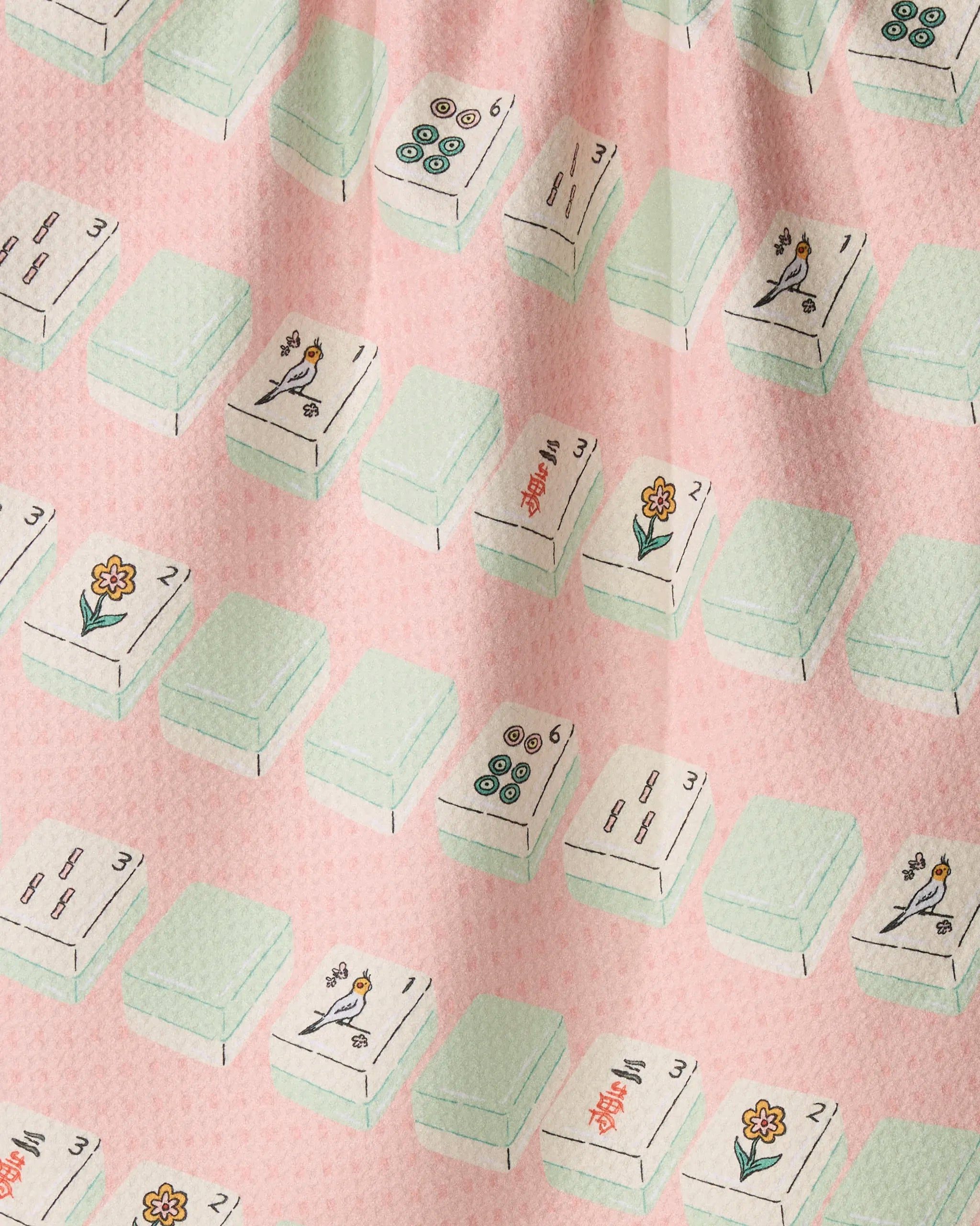 Tile Parade by Geometry is a patterned fabric showcasing rows of Mahjong tiles with diverse designs—flowers, circles, and birds—set against a light pink background. Made from recycled materials, this fabric features subtle folds and a textured surface.