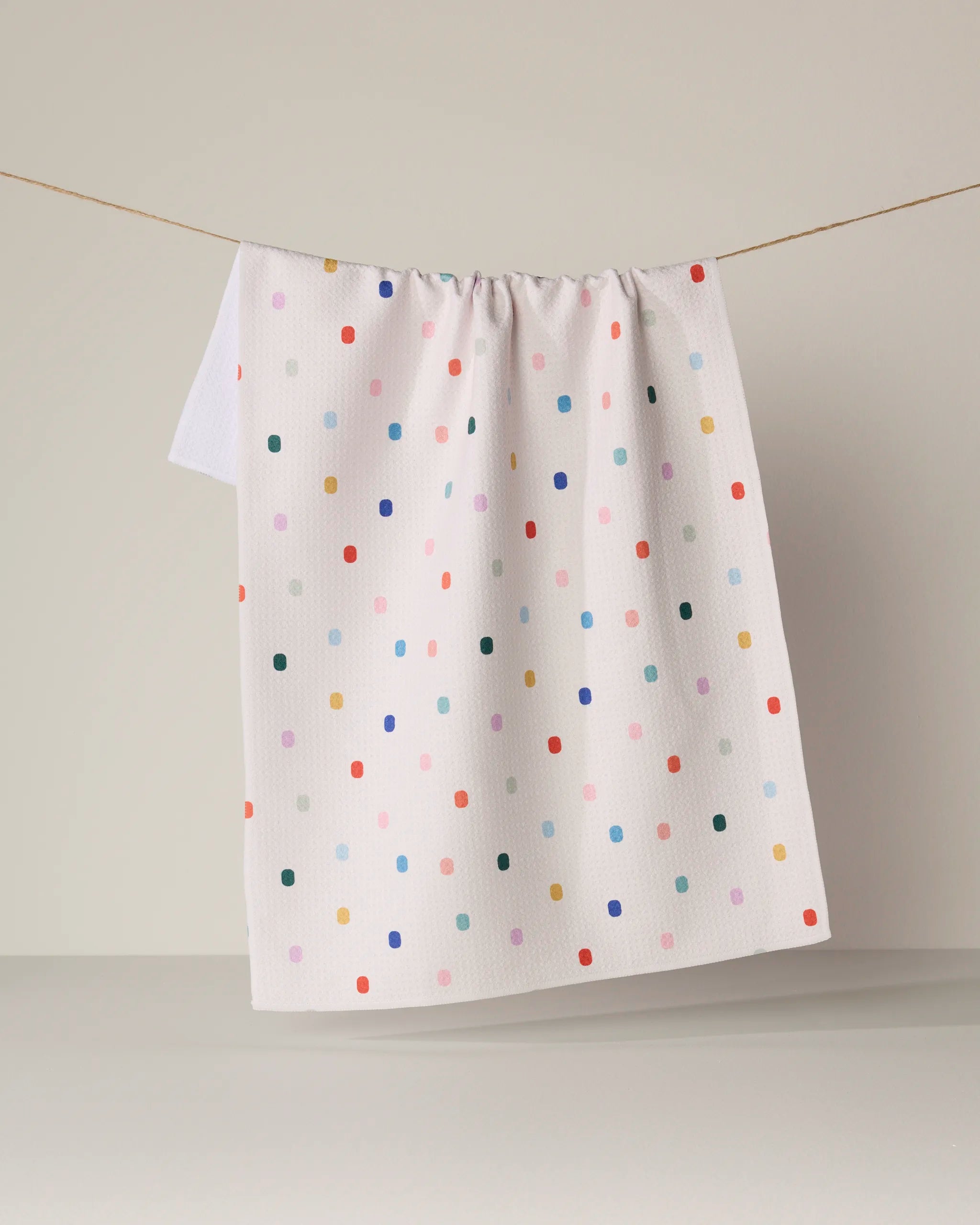 A Geometry Tile Shuffle tea towel, featuring a colorful polka-dot pattern and made from recycled materials, hangs from a string against a neutral background.