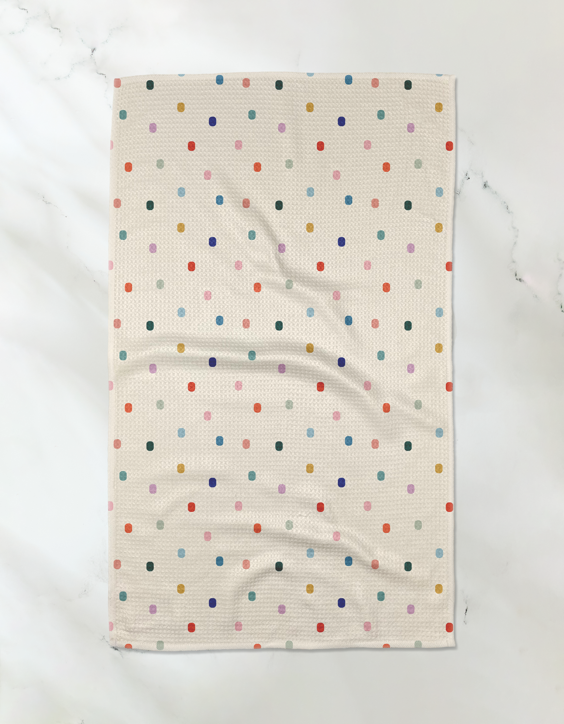 A Geometry "Tile Shuffle" recycled kitchen towel, featuring a grid of colorful polka dots in red, blue, green, orange, and pink on a beige background, is laid flat on a white marble surface.