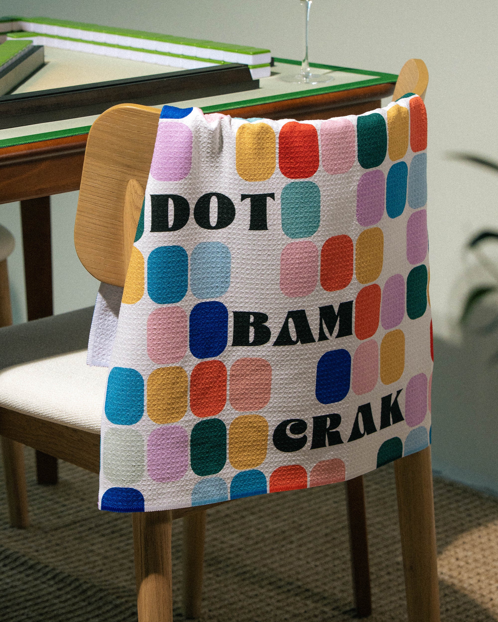A vibrant Geometry Tile Talk Tea Towel, adorned with rounded squares and the words DOT BAM CRAK, is draped over a wooden chair beside a table set with books and a wine glass. This eco-friendly tea towel from Geometry adds practical flair and vivid style to any space.