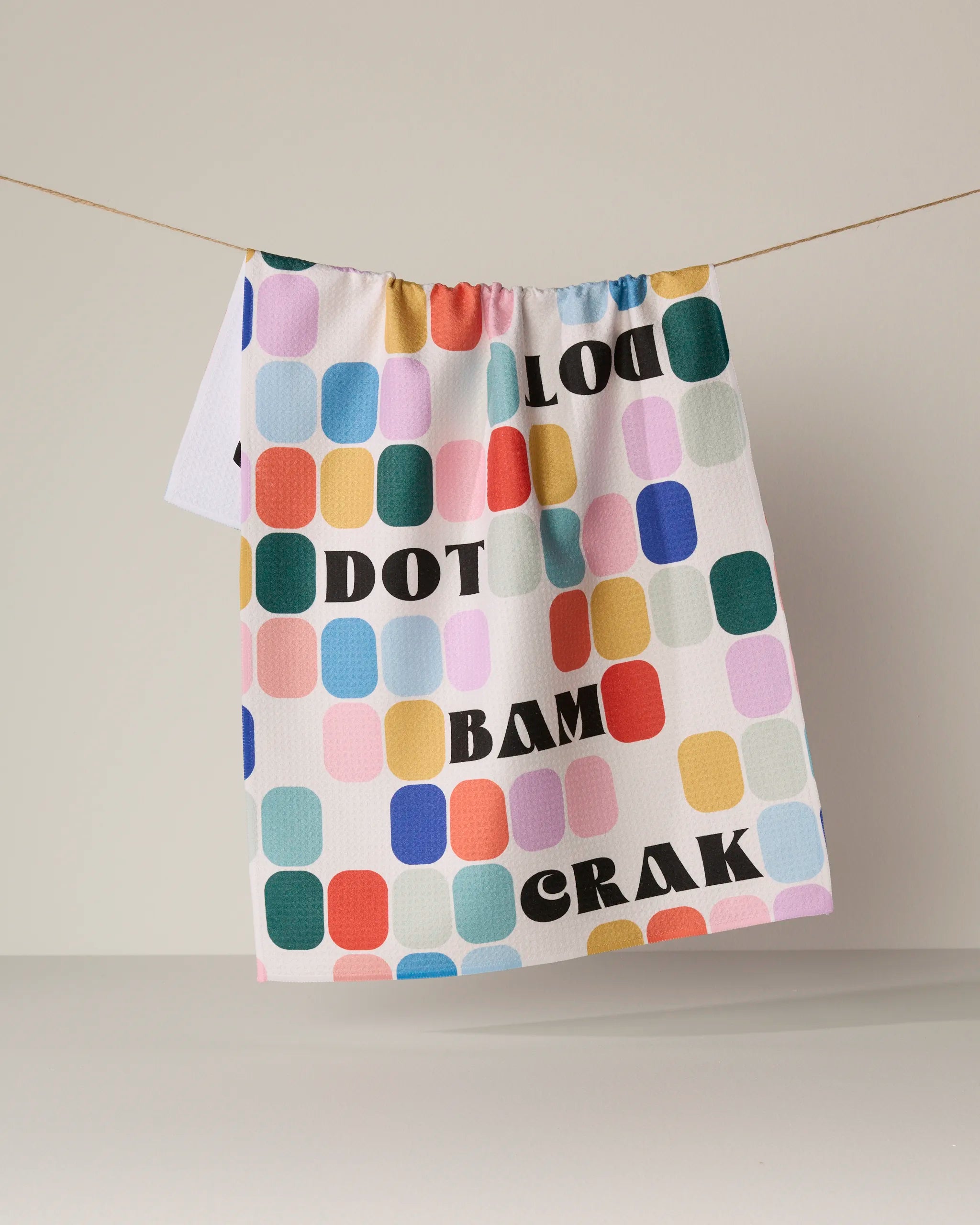 A vibrant tea towel from Geometry called "Tile Talk," featuring rounded squares in pastel hues and crafted from recycled materials, hangs on a string. Bold black letters on the towel spell out LOOK DOT BAM CRAK against a plain, neutral background.