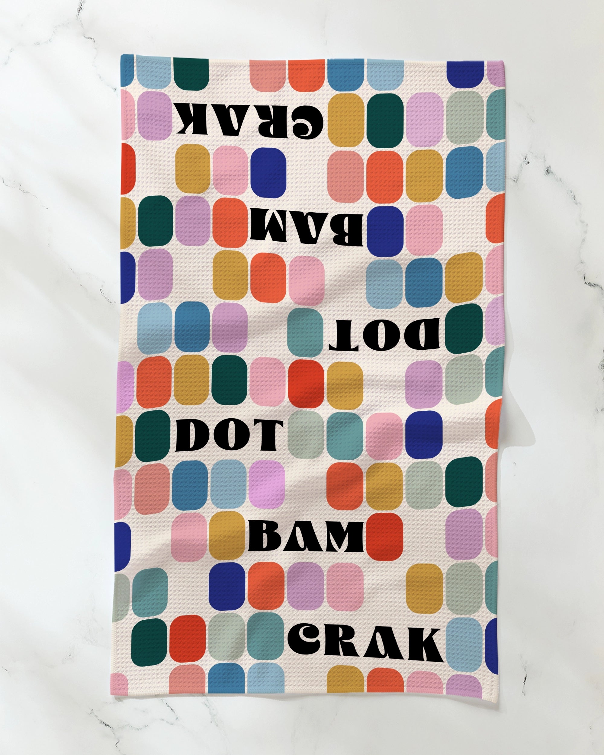 A vibrant Geometry Tile Talk Tea Towel features a grid of rounded squares in an array of colors and bold black text reading DOT BAM CRAK—mirrored upside down for extra flair—bringing playful charm to your kitchen. Ultra-absorbent and stylish, this Geometry towel is displayed on a white marble surface, making it the perfect blend of form and function for your space.