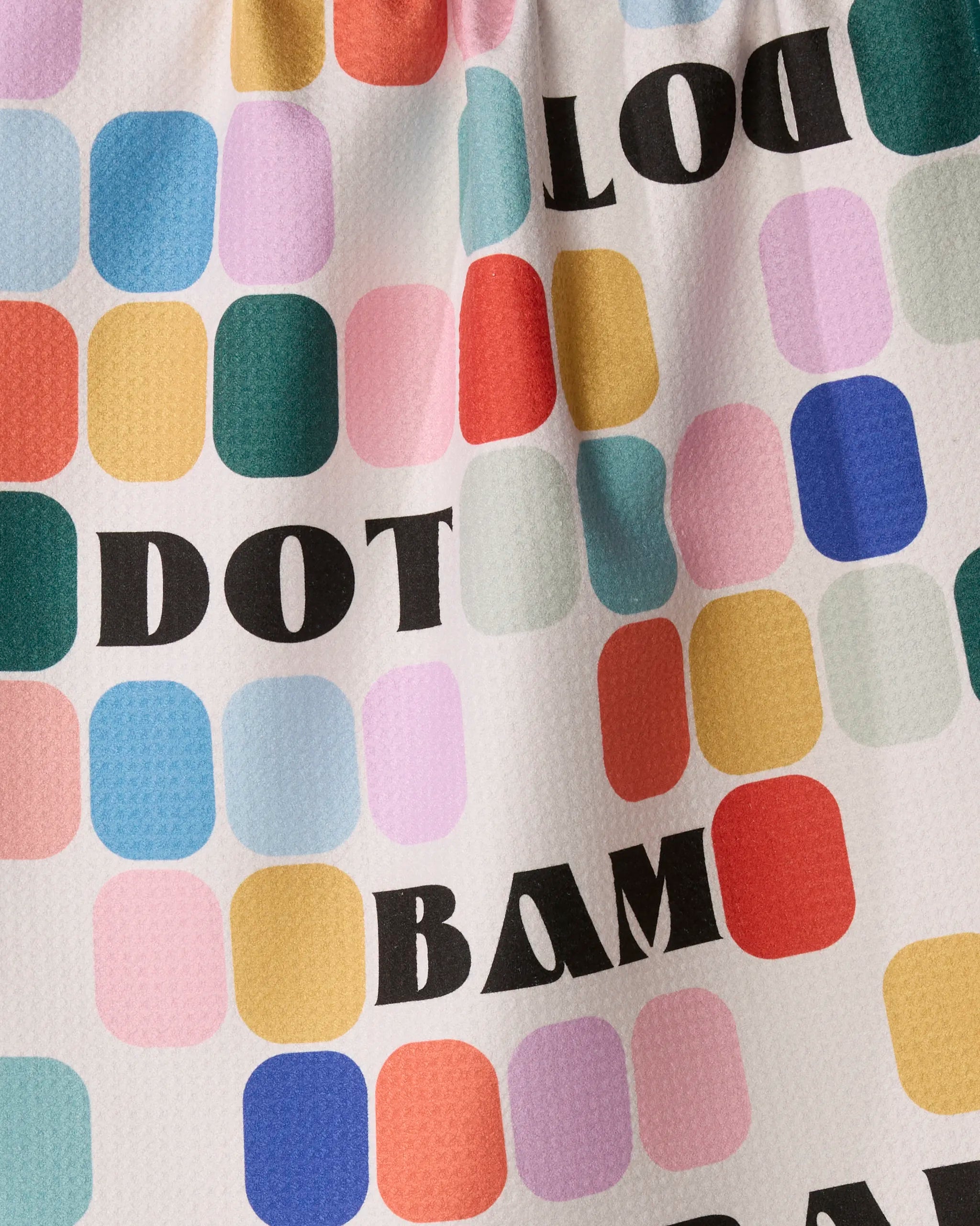 Colorful squares and rectangles in various hues form a patterned background with bold black text reading “Tile Talk” printed across the textured tea towel by Geometry, which is crafted from recycled materials.