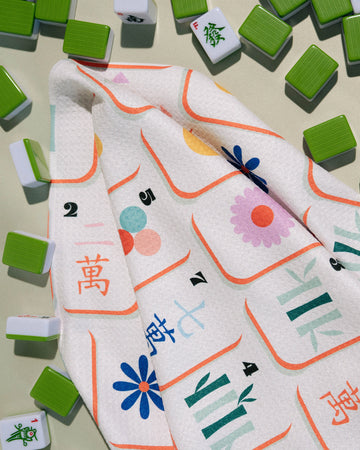 The Geometry Tile Toss kitchen tea towel, featuring a vibrant floral pattern and Chinese characters on a white background, rests among scattered green and white Mahjong tiles on a light surface.
