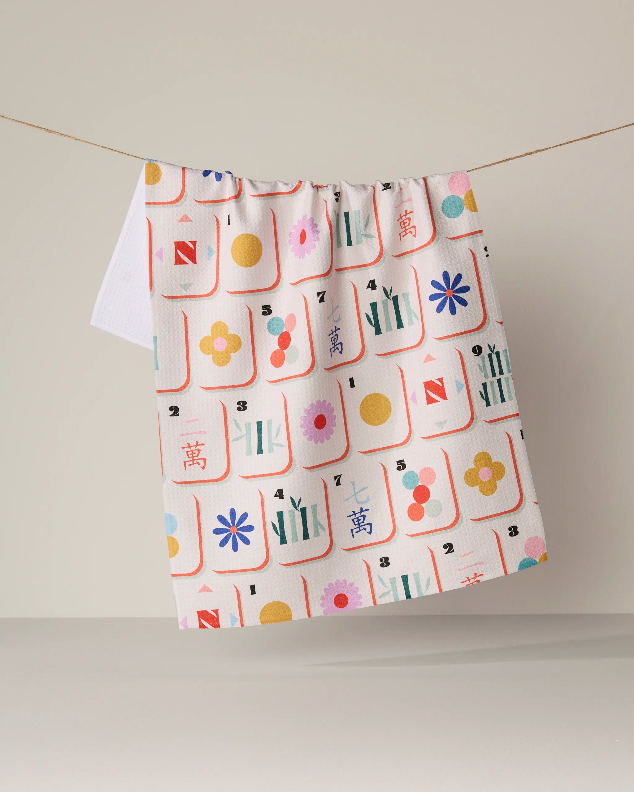 A kitchen towel from Geometry, called "Tile Toss," made from white recycled material and featuring colorful mahjong tile patterns with flowers, circles, and bamboo, hangs on a string against a neutral background.