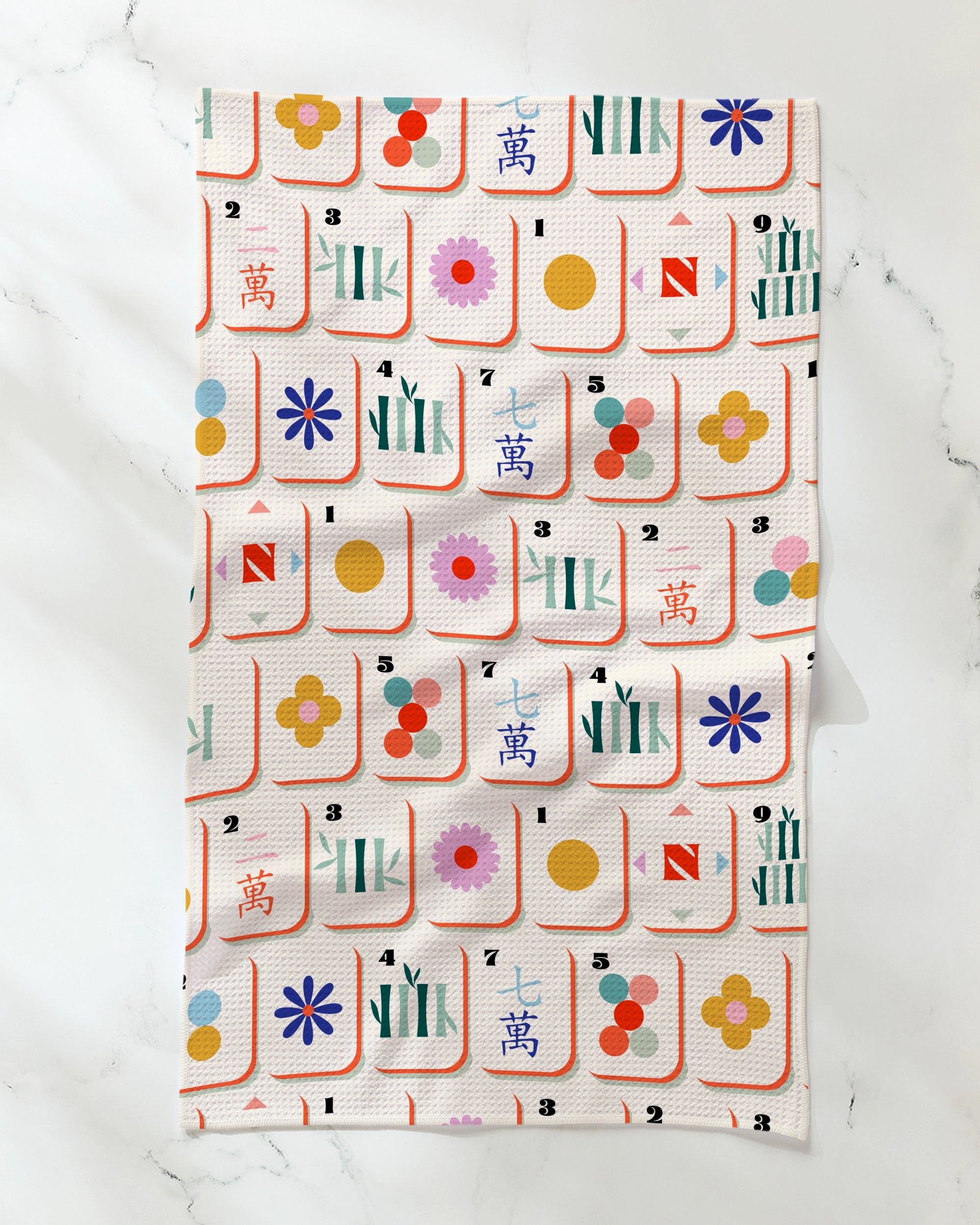 The Geometry "Tile Toss" kitchen tea towel, crafted from recycled materials, showcases a vibrant Mahjong tile-inspired pattern with stylized circles, bamboo, and character designs on a crisp white background and is displayed flat on a white marble surface.