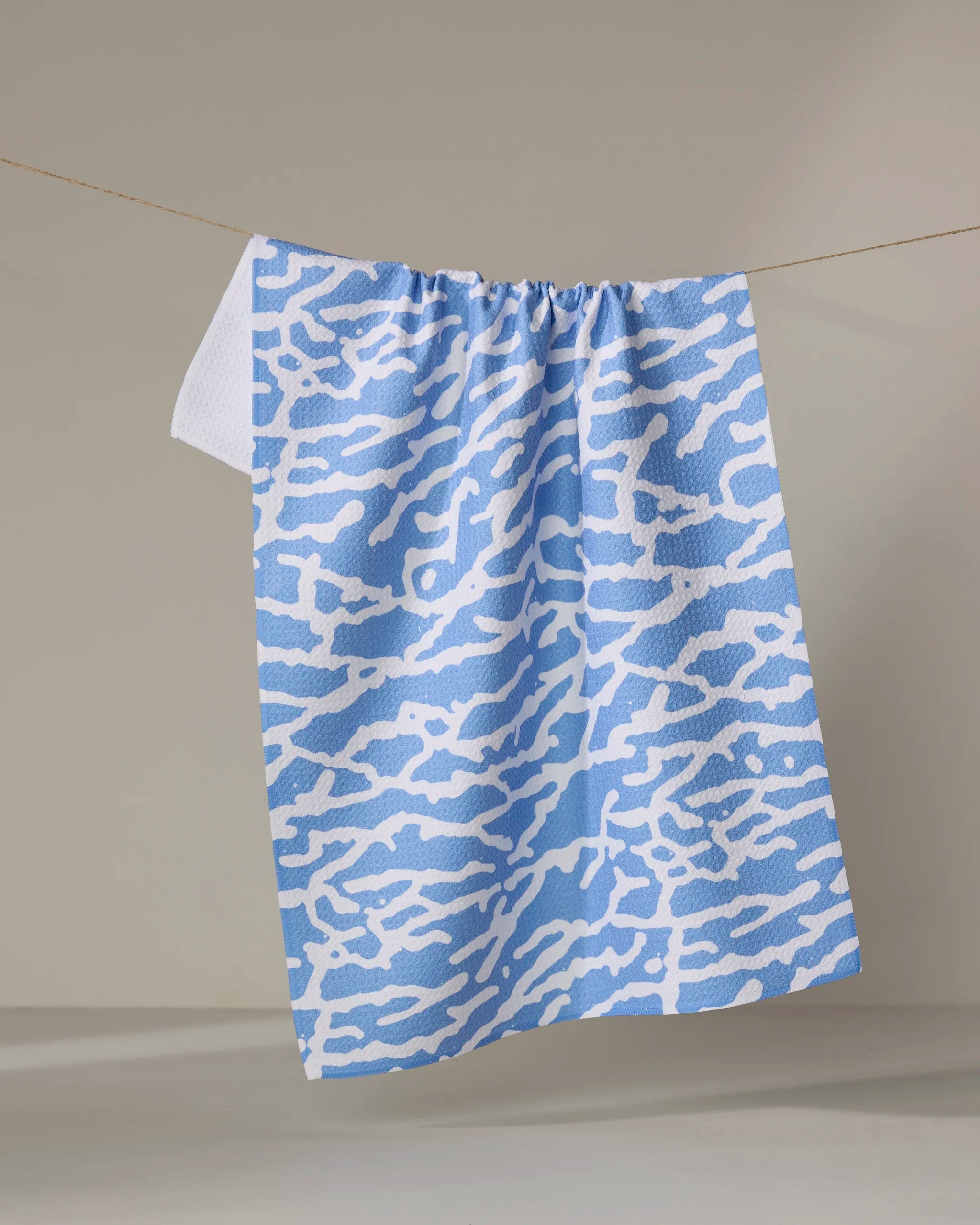 The Tinker kitchen towel by Sissy Moon, featuring a blue and white abstract wavy design, hangs on a thin clothesline against a neutral background.