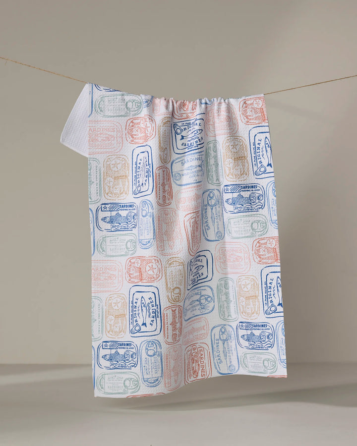 The "Tinned Sardines" dish towel by Hufton Studio features a vintage-inspired sardine can label pattern in blue, red, and green on white, shown hanging on a line against a neutral background.