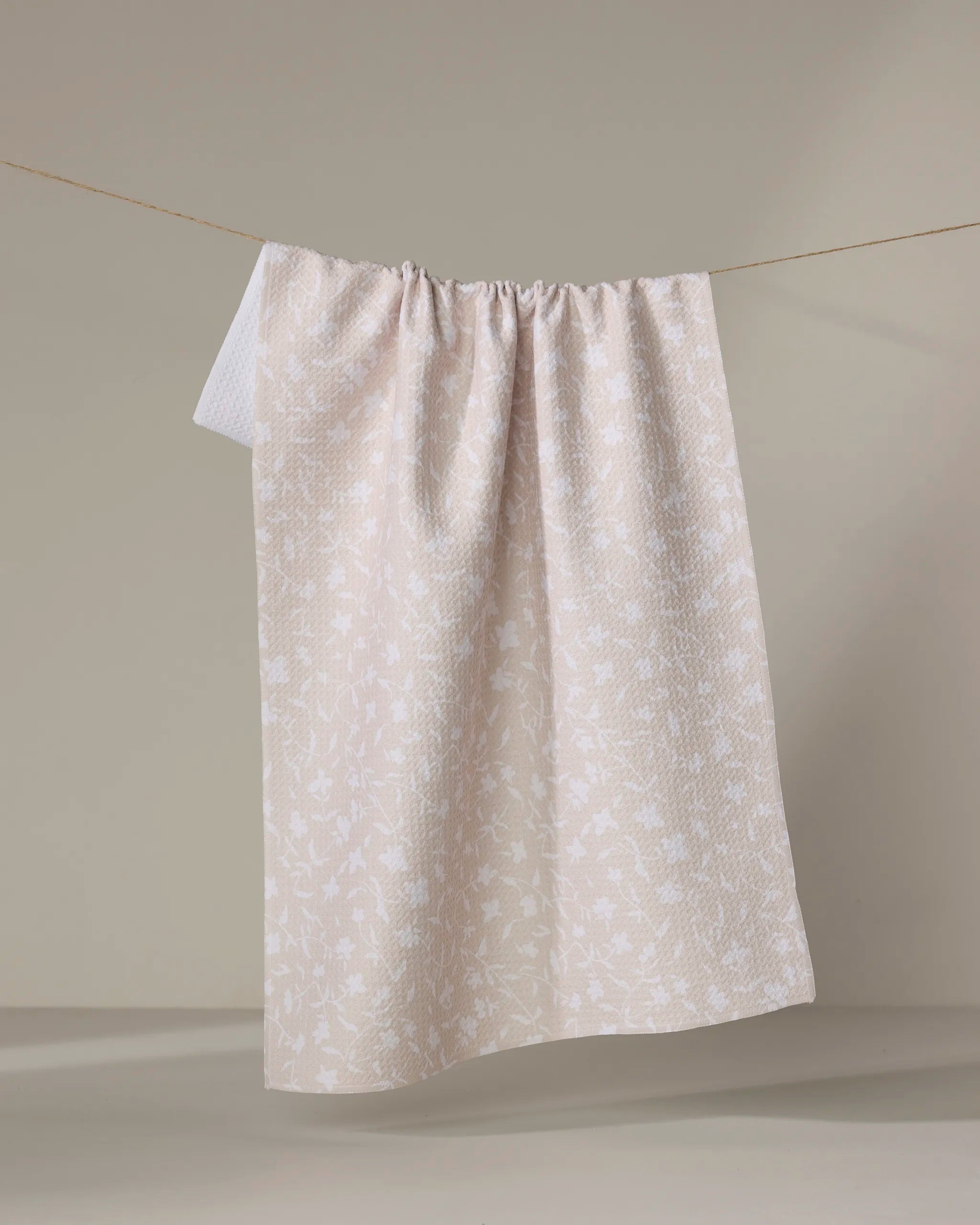 The Tiny Floral tea towel by Aubrey Fairchild, featuring a cream color with a subtle white floral pattern, is shown draped over a string against a plain beige background.
