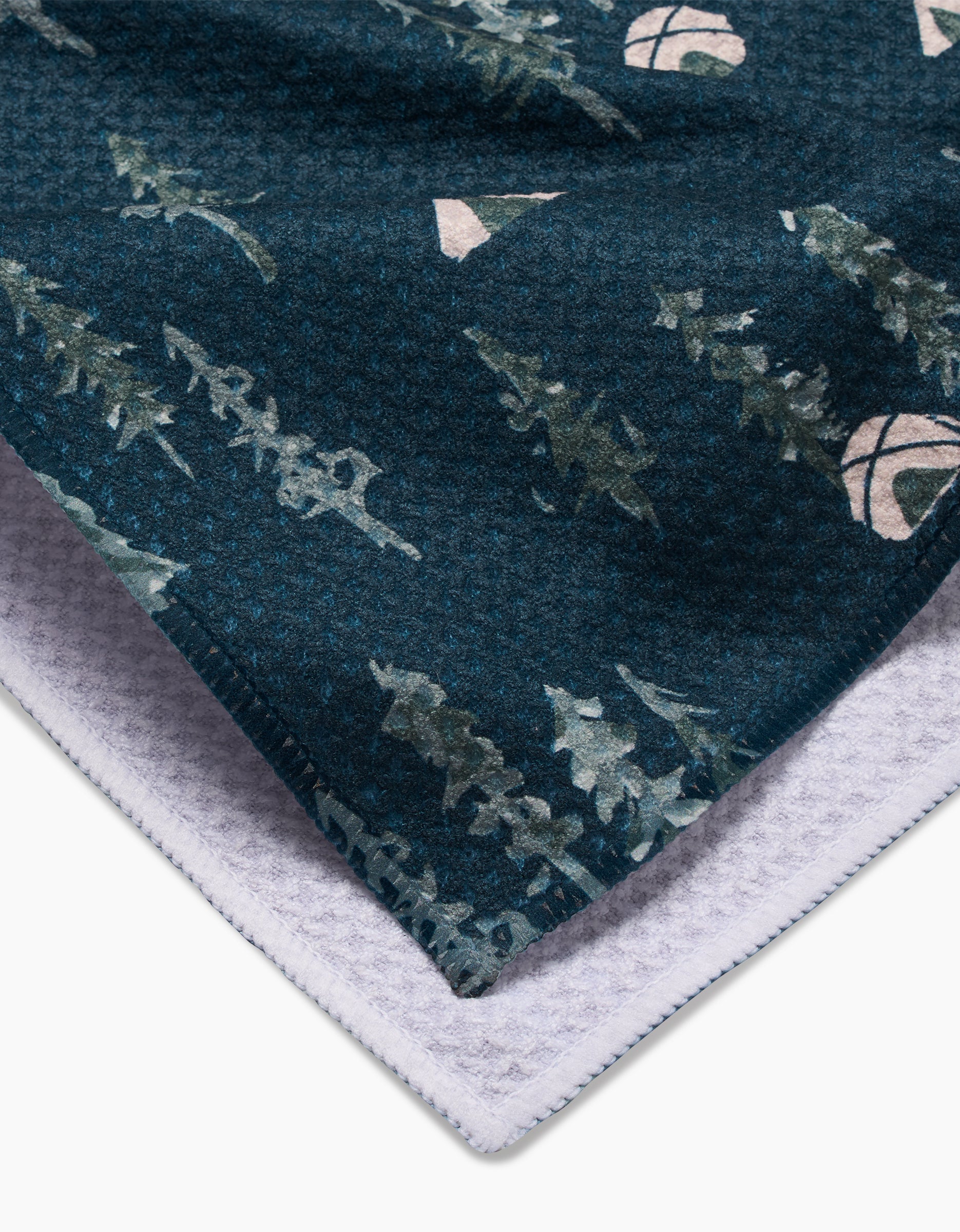 A close-up of Twig + Spark’s Tiny Tents Geoweave fabric shows its blue-green forest and mountain print, ultra-absorbent texture, soft white underside, and a folded corner revealing its layered construction.