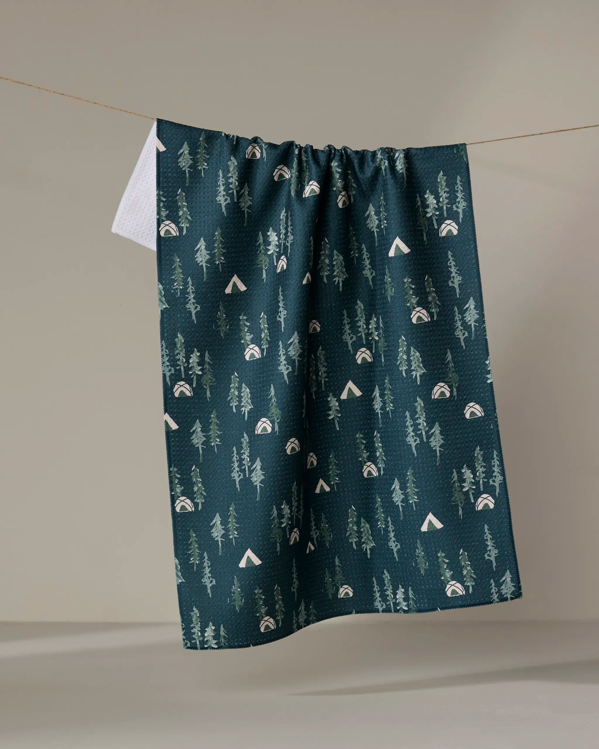 The Twig + Spark Tiny Tents tea towel, made from ultra-absorbent recycled materials, features a dark green design with trees, rainbows, and white triangles. It hangs on a clothesline against a plain, light background.