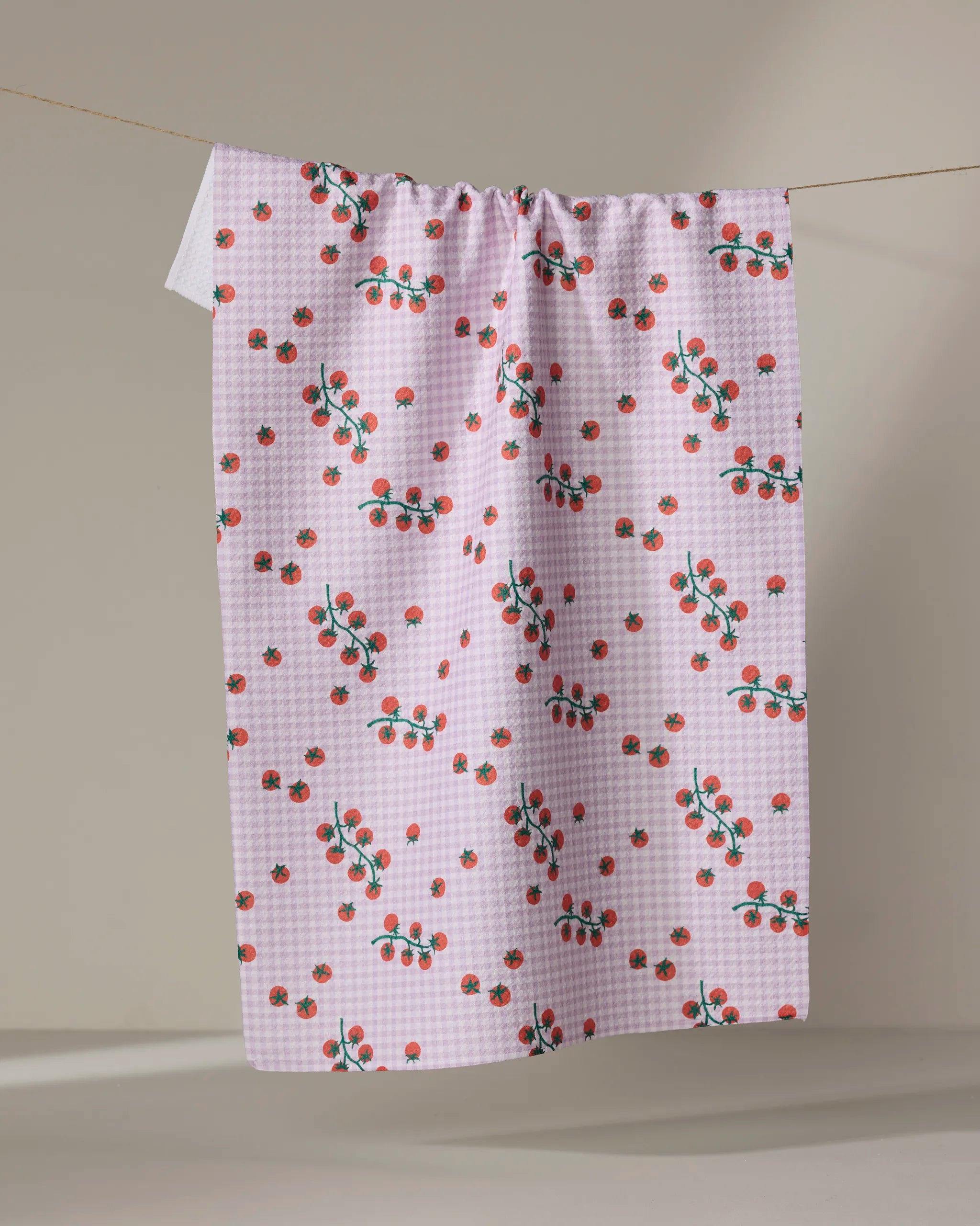 The Tomato Gingham kitchen towel by Brittany Jepsen features pink and white checks with red cherries and green leaves, hanging on a clothesline against a neutral background.