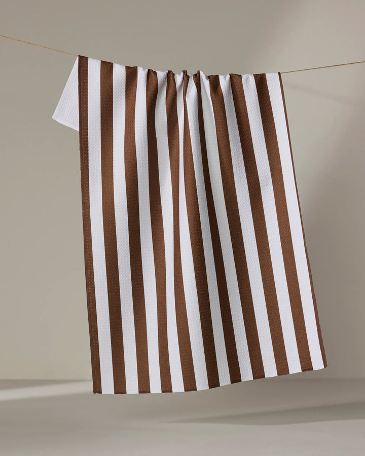 The Kailee Wright Toni Stripe tea towel, featuring white and brown vertical stripes and made from recycled materials, hangs on a string against a neutral background.