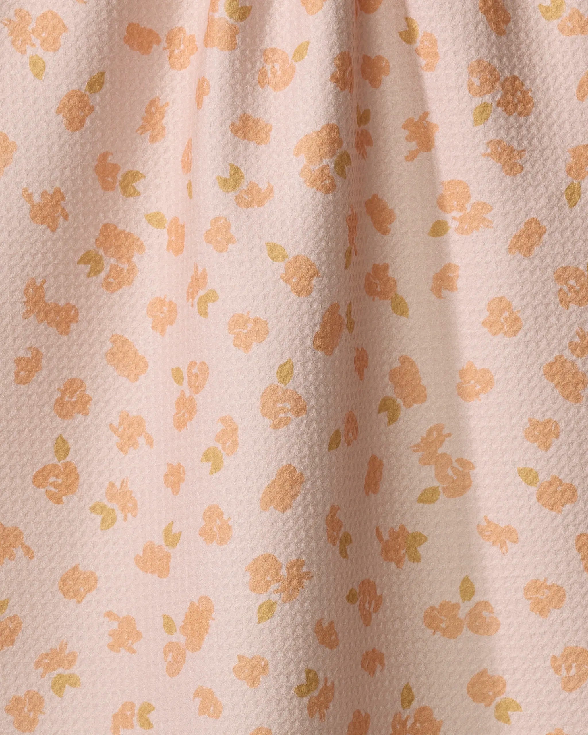 The Tossed Wildflowers Pink tea towel by Mandy Arroyo features a light pink, textured design with scattered peach and yellow floral prints and green leaves. Made from ultra-absorbent recycled materials, it has gentle folds at the top.