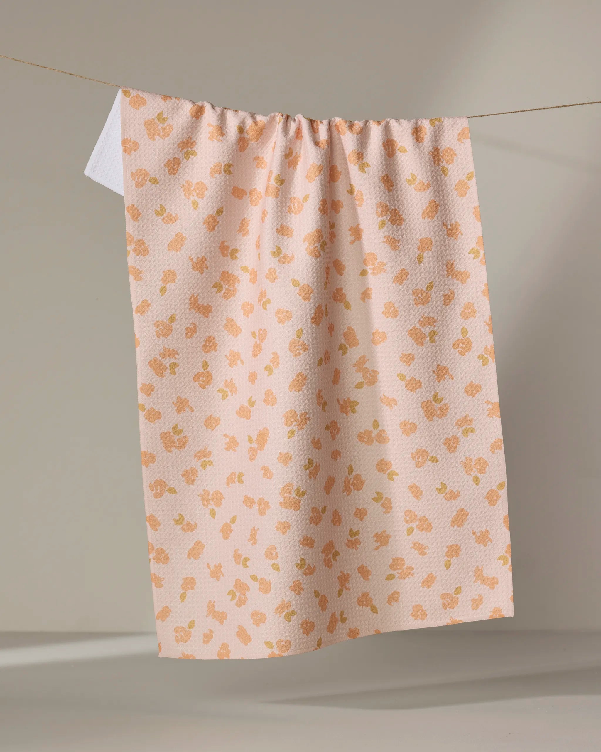 The Tossed Wildflowers Pink tea towel by Mandy Arroyo features a peach hue with orange and yellow floral motifs. Made from recycled materials, it hangs on a line against a neutral background.