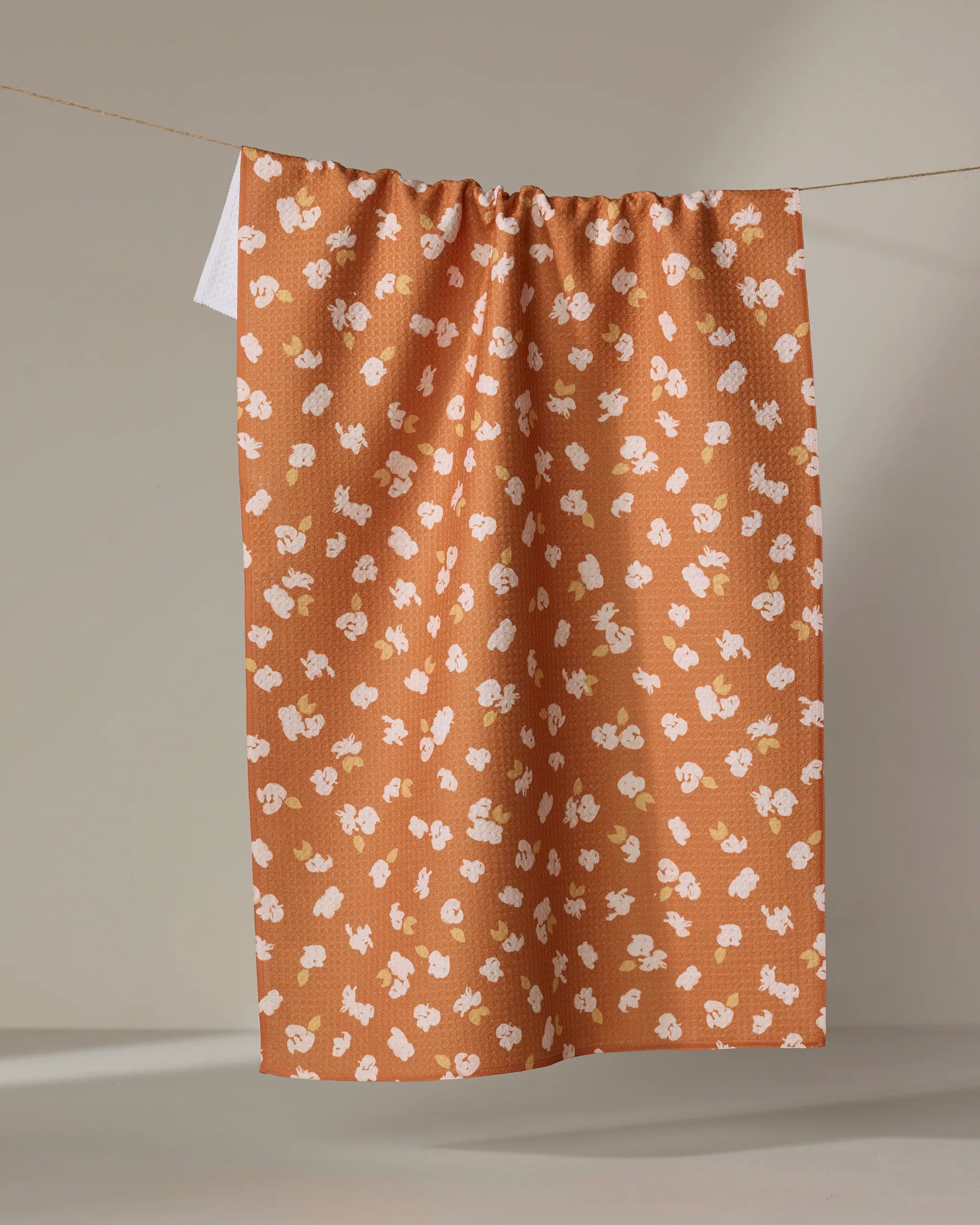 The Tossed Wildflowers Rust tea towel by Mandy Arroyo features a rust-orange color, a scattered wildflower print in white, pink, and yellow, and is made from recycled materials, shown hanging vertically against a neutral background.