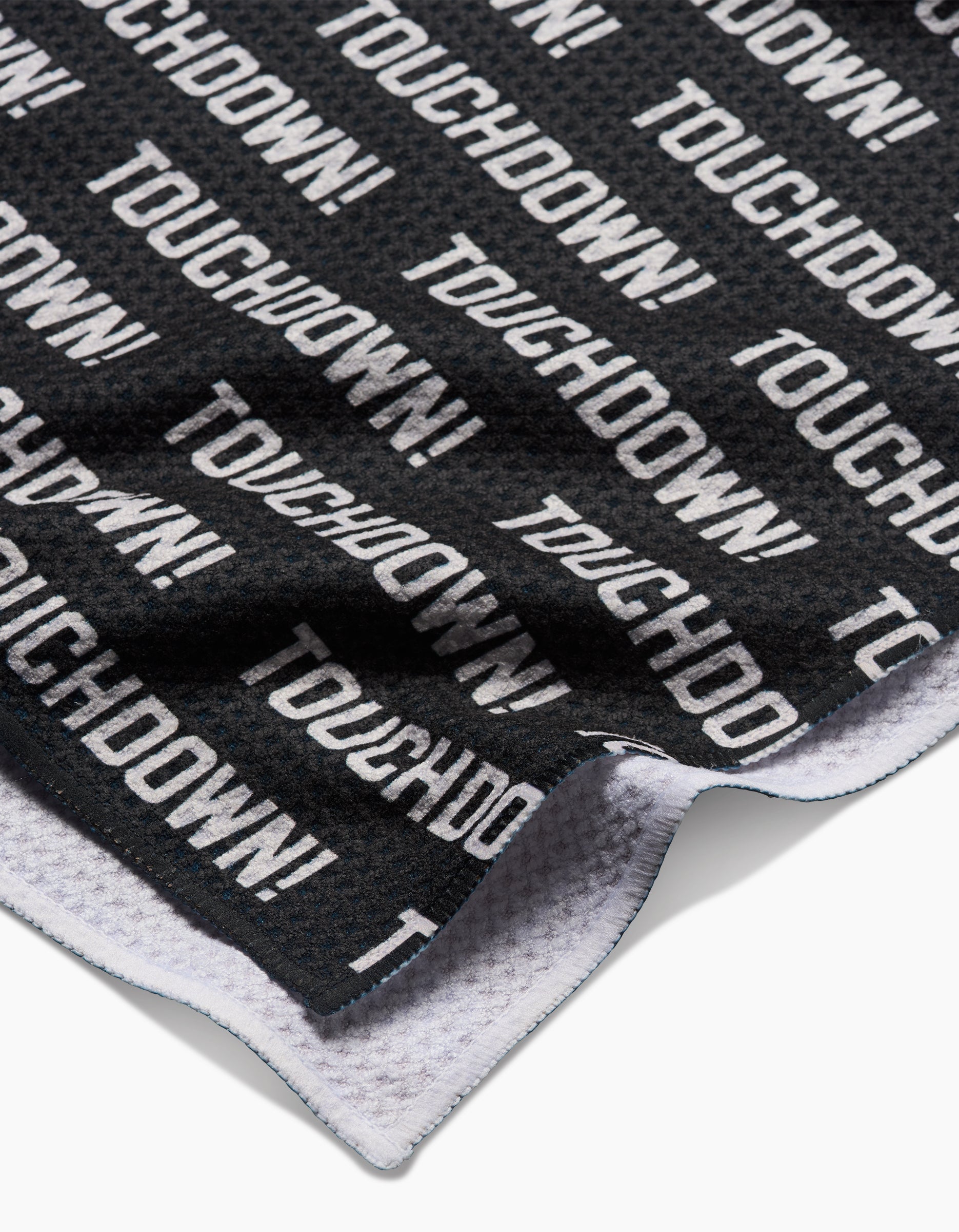 A close-up of a black GEOMETRY Geoweave Touchdown Tea Towel shows TOUCHDOWN! in bold white letters. The edge is slightly folded to reveal the white underside, and its made from recycled materials.