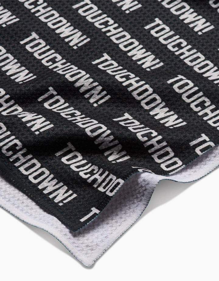A close-up of a black GEOMETRY Geoweave Touchdown Tea Towel shows TOUCHDOWN! in bold white letters. The edge is slightly folded to reveal the white underside, and its made from recycled materials.