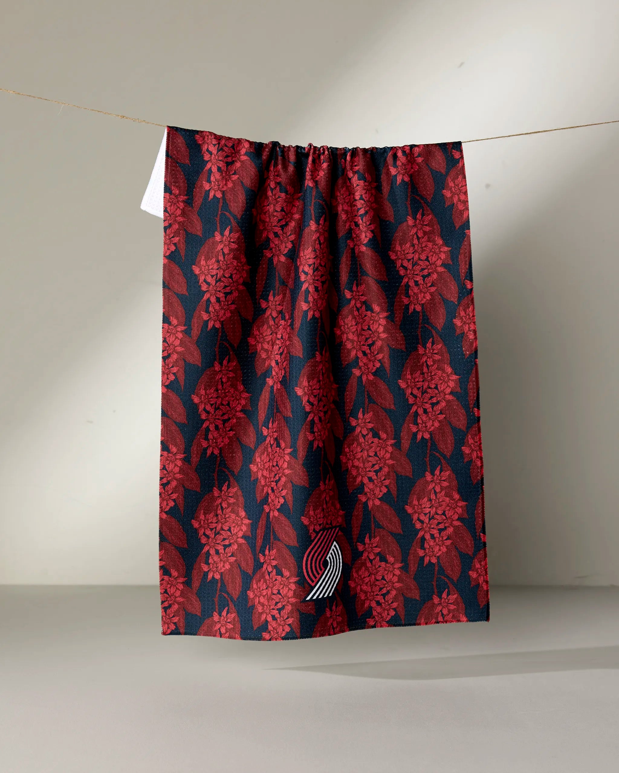 A dark Trail Blazers Floral tea towel by NBALAB x Geometry with a red floral pattern hangs on a string against a plain background, featuring a small white curved logo near the bottom edge.