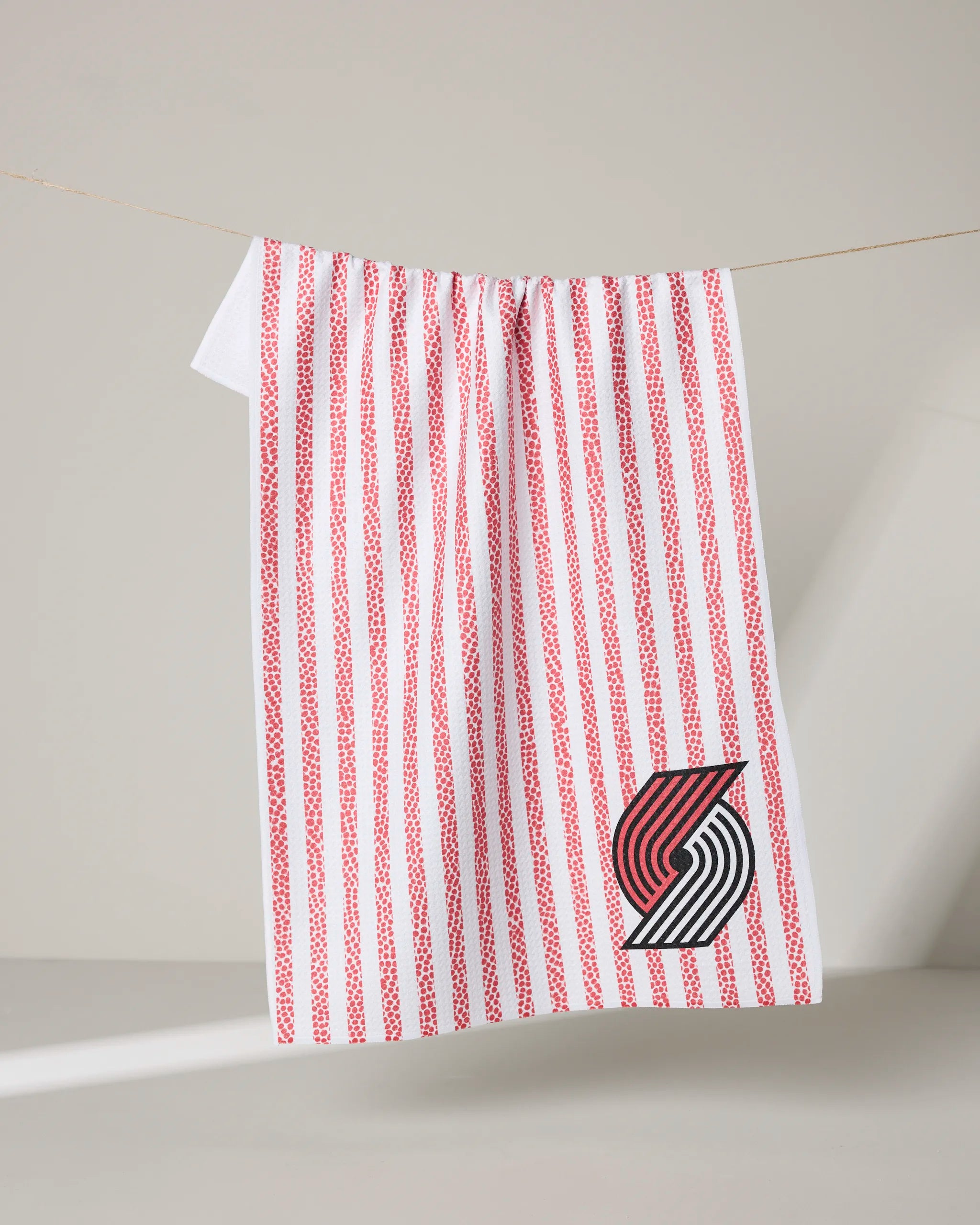 A white NBALAB x Geometry Trail Blazers Grip Stripe tea towel with vertical red stripes hangs on a clothesline, featuring the Portland Trail Blazers logo in black, red, and white at the bottom right corner.