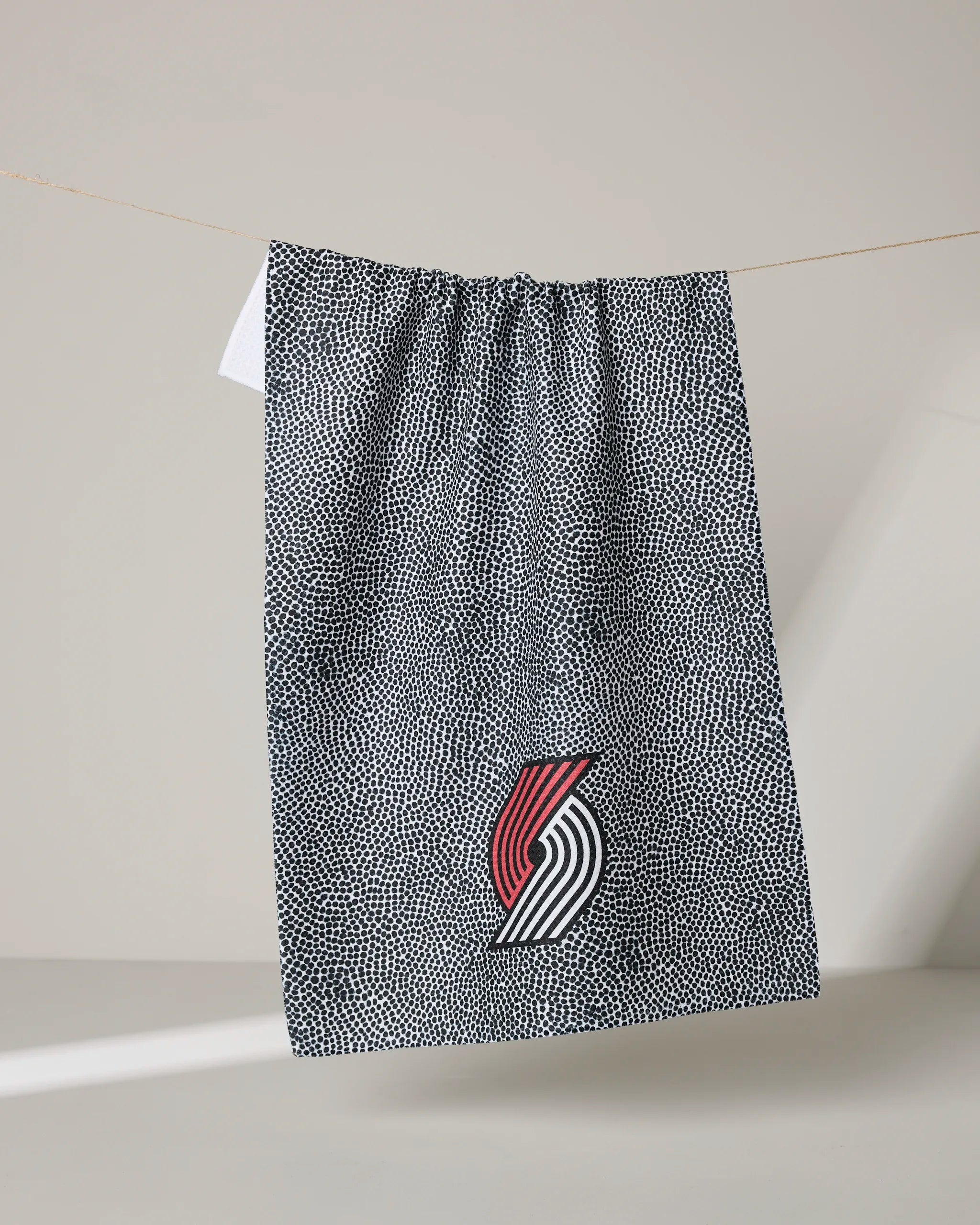 A black and white speckled NBALAB x Geometry Trail Blazers Grip tea towel hangs on a line, featuring the Portland Trail Blazers logo with red, white, and black lines near the bottom center.