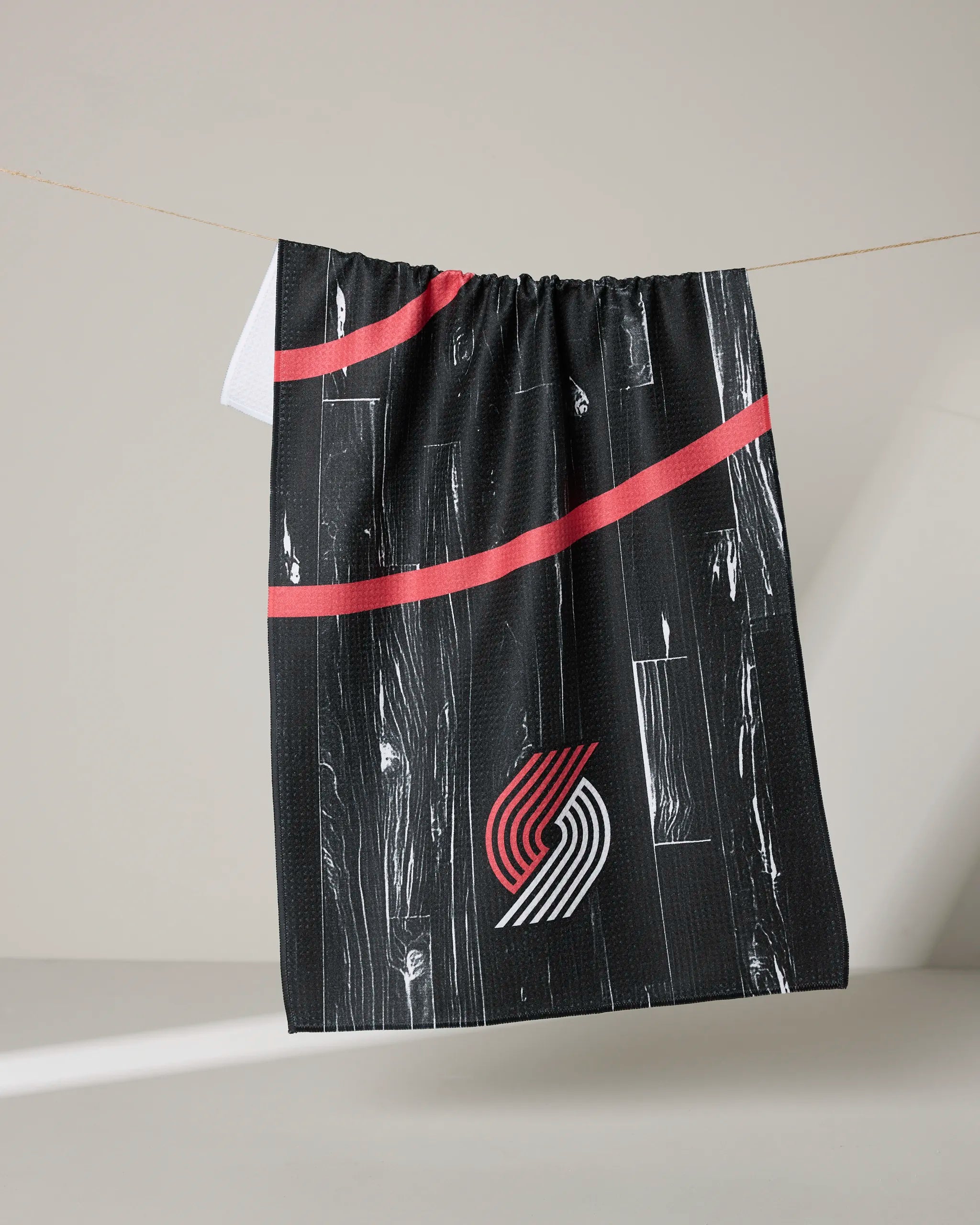 The NBALAB x Geometry Trail Blazers Zone tea towel, featuring a white and red abstract woodgrain design with the Portland Trail Blazers logo, hangs on a clothesline against a neutral background.